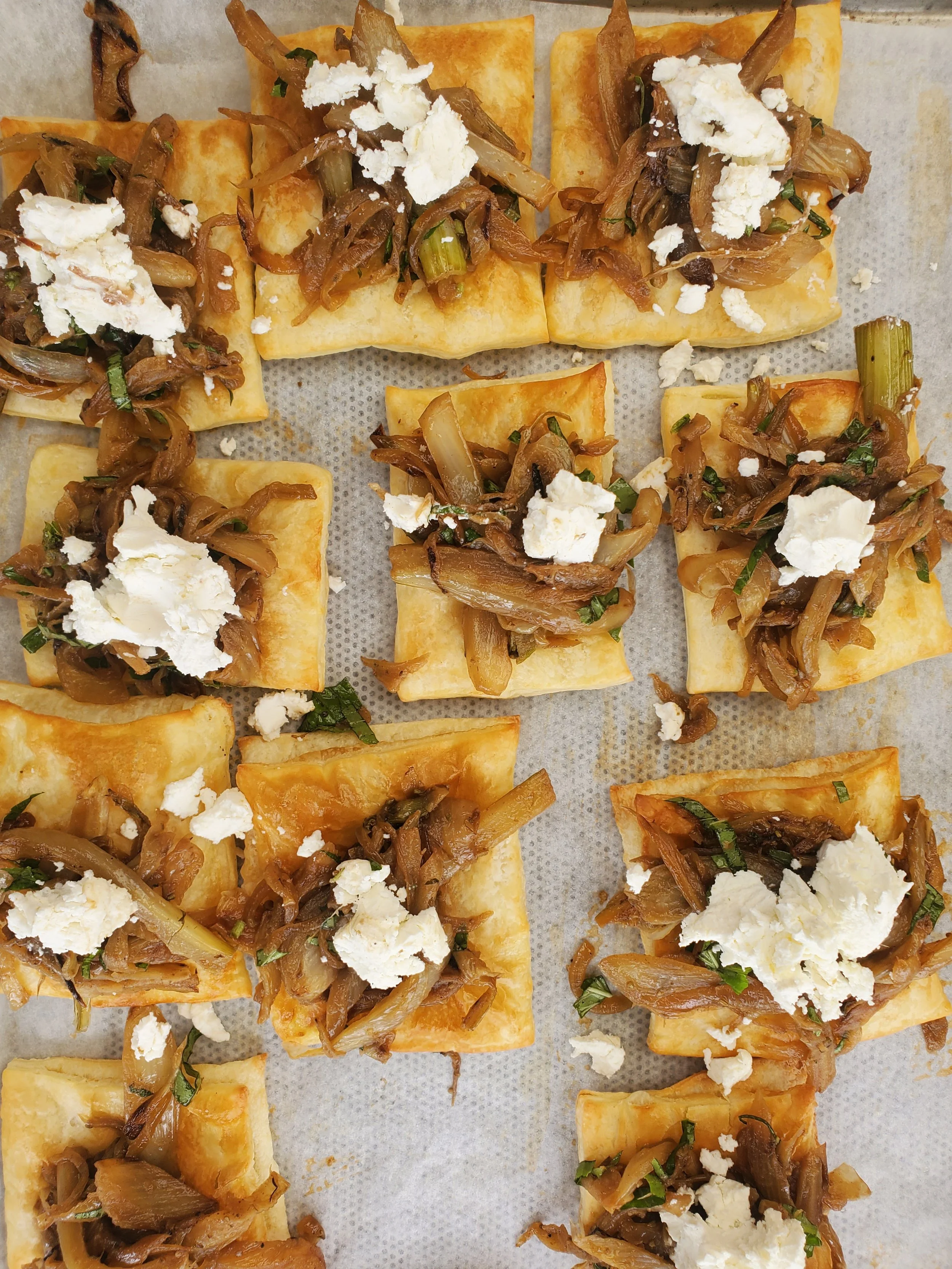 Multiple rectangular pieces of baked pastry topped with cooked onions, crumbled white cheese, and chopped herbs, arranged on parchment paper.