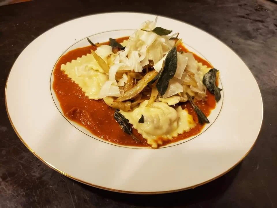A plate of ravioli pasta topped with sautéed vegetables and garnished with shredded cheese in tomato sauce.