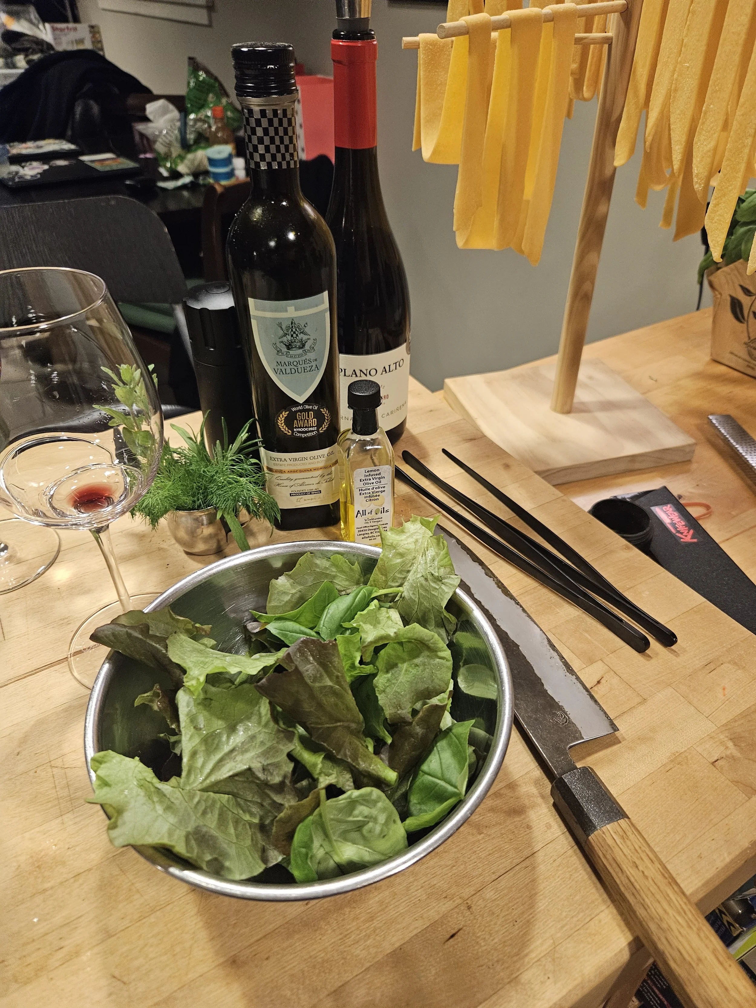 A kitchen table with a bowl of green leafy lettuce, bottles of wine, a small bottle of olive oil, a butter knife, a pair of chopsticks, and a cutting board with yellow pasta hanging on a drying rack.