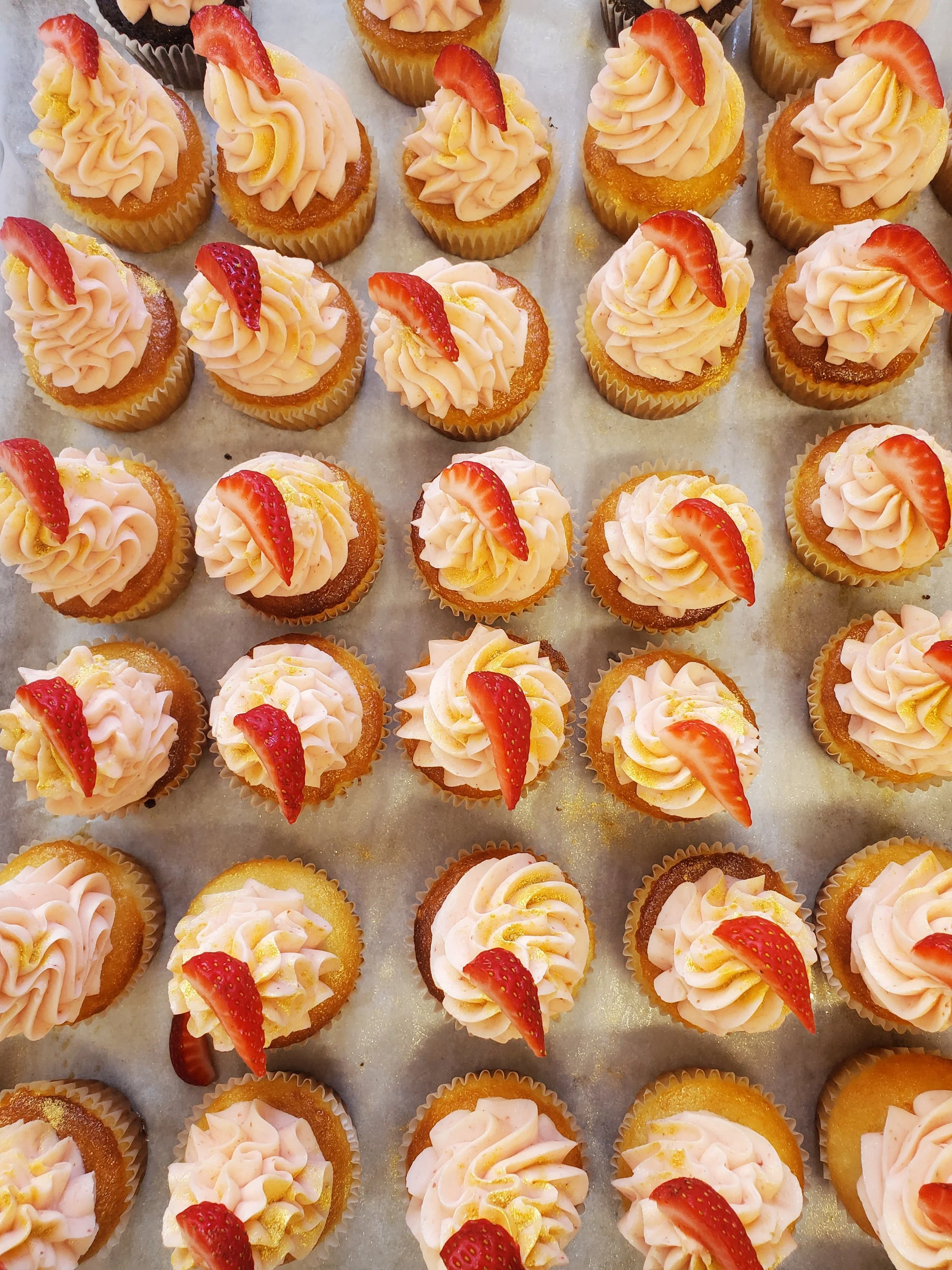 Numerous small cupcakes with pink frosting, topped with a slice of strawberry and yellow sprinkles, arranged on a baking sheet.