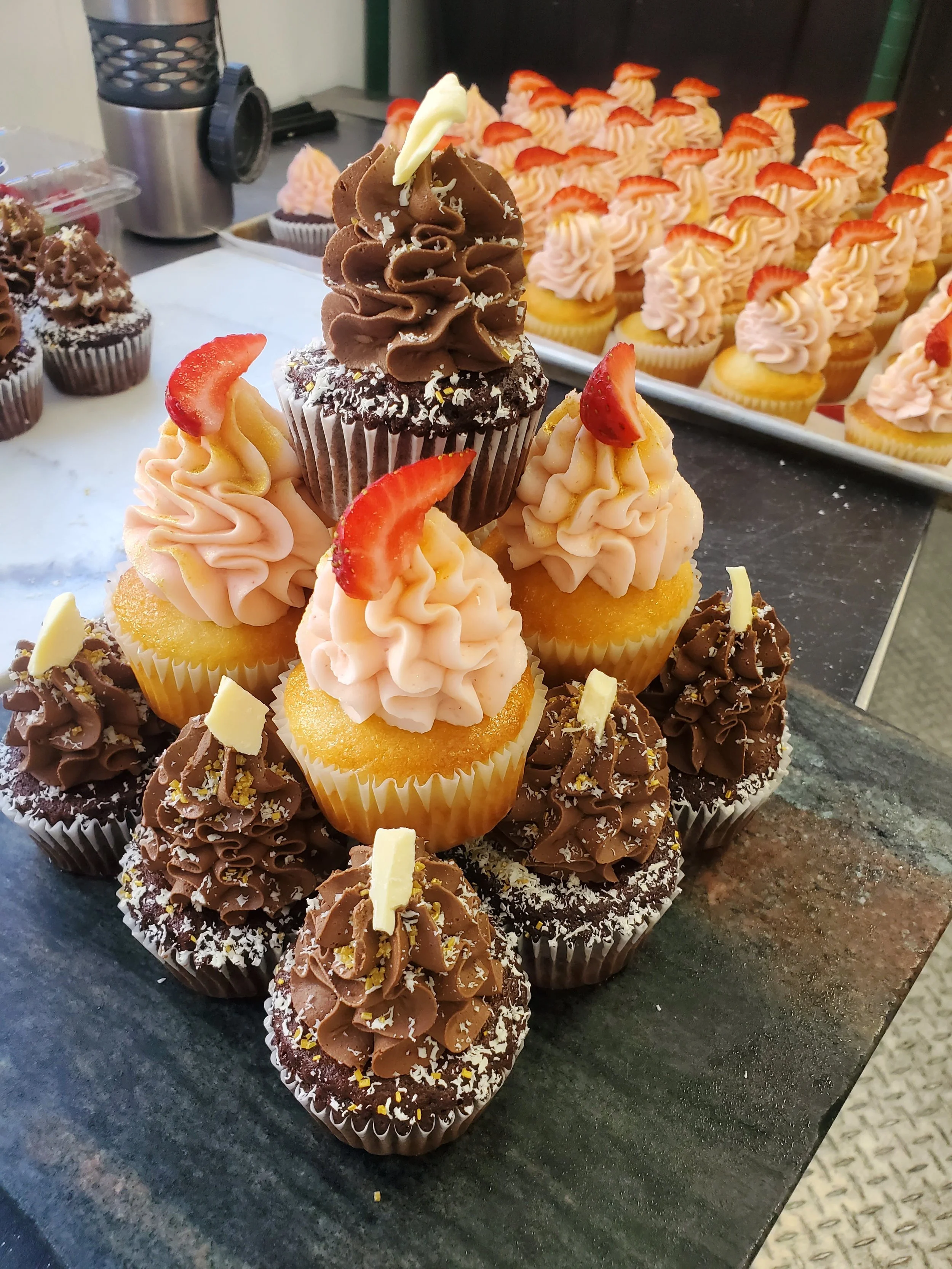 Assorted cupcakes on a dark surface, some topped with strawberries and white chocolate shavings, others with chocolate frosting and sprinkles, with a tray of pink frosted cupcakes in the background.