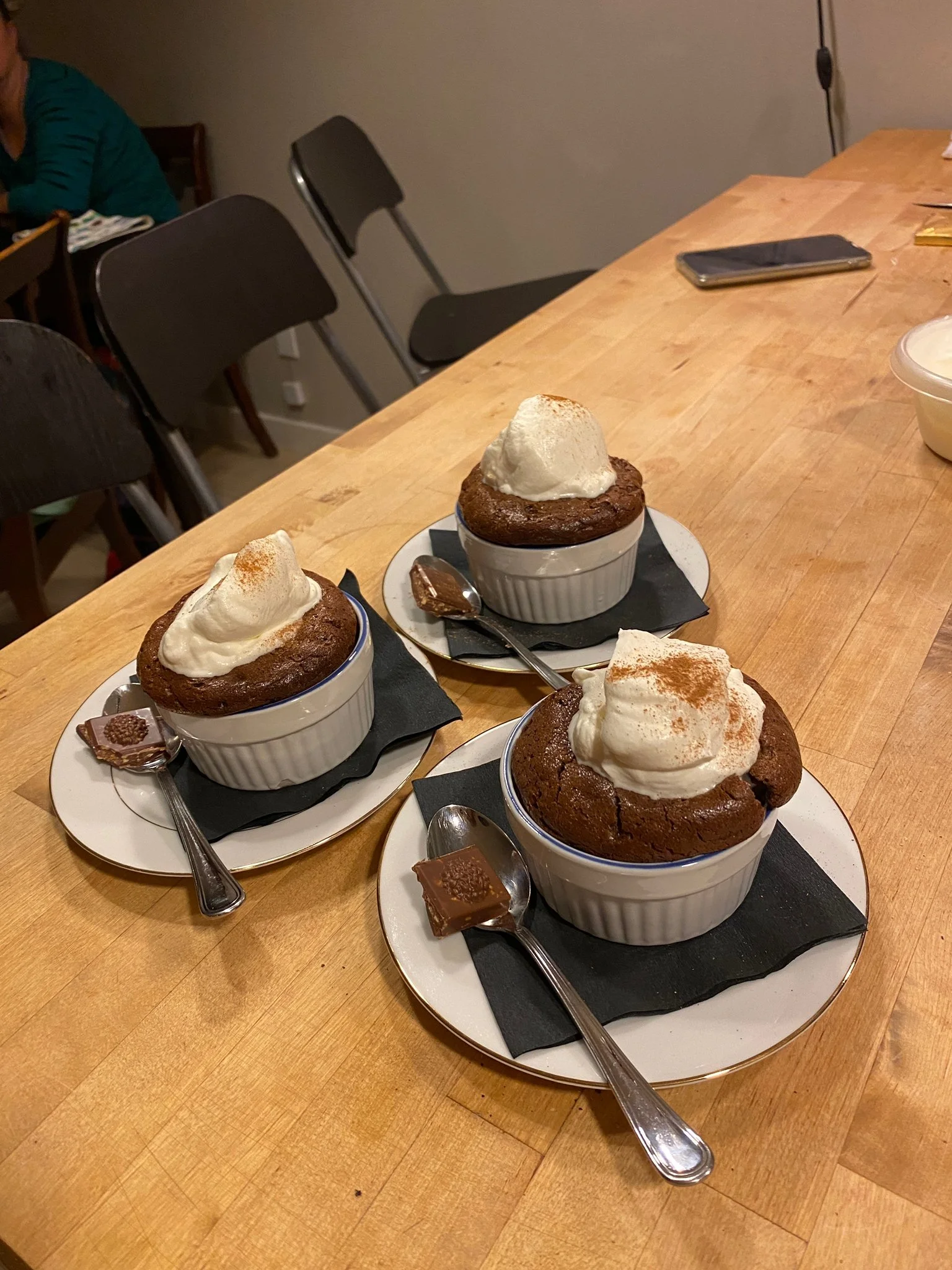 Three chocolate lava cakes topped with whipped cream and cinnamon powder, each served in a small white ramekin on black napkins with spoons on white oval plates, placed on a wooden table.