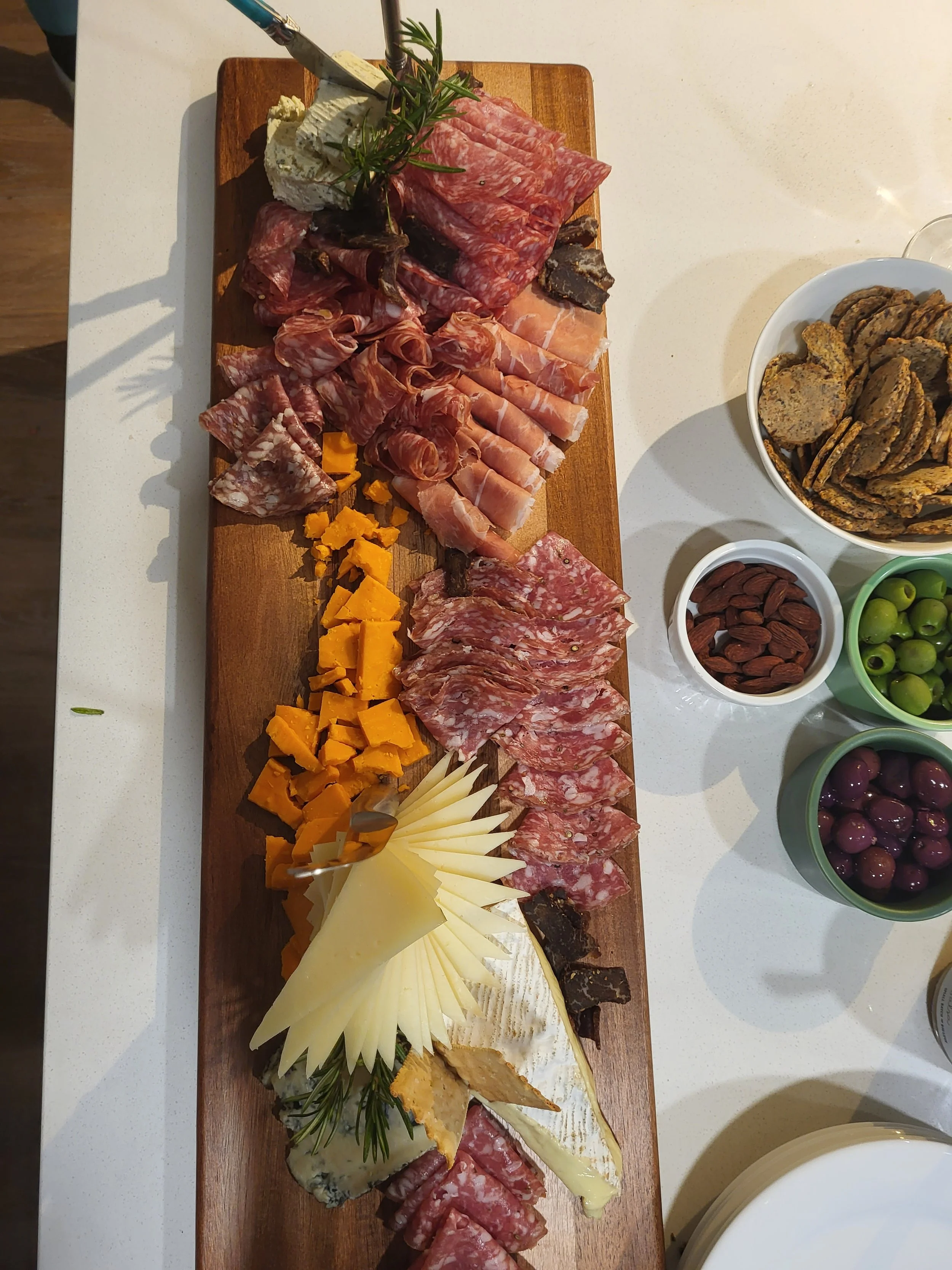A wooden serving board with an assortment of sliced cured meats and cheeses, garnished with rosemary and slices of cheddar cheese, with bowls of crackers, almonds, green olives, and purple olives on a white countertop.