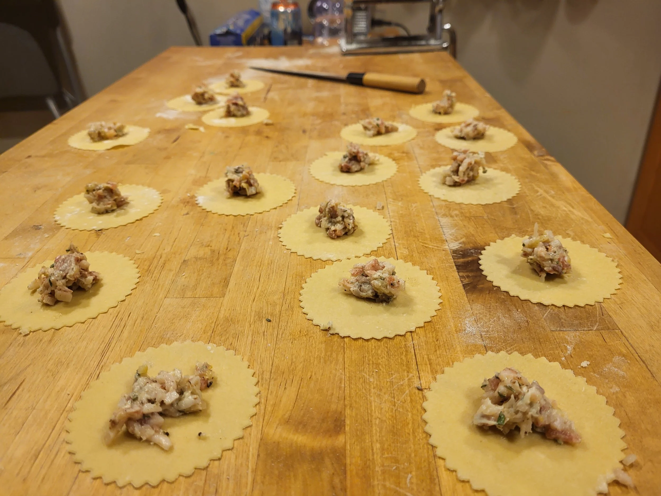 Uncooked ravioli pasta shells filled with meat or cheese on a wooden countertop, with a knife and kitchen supplies in the background.