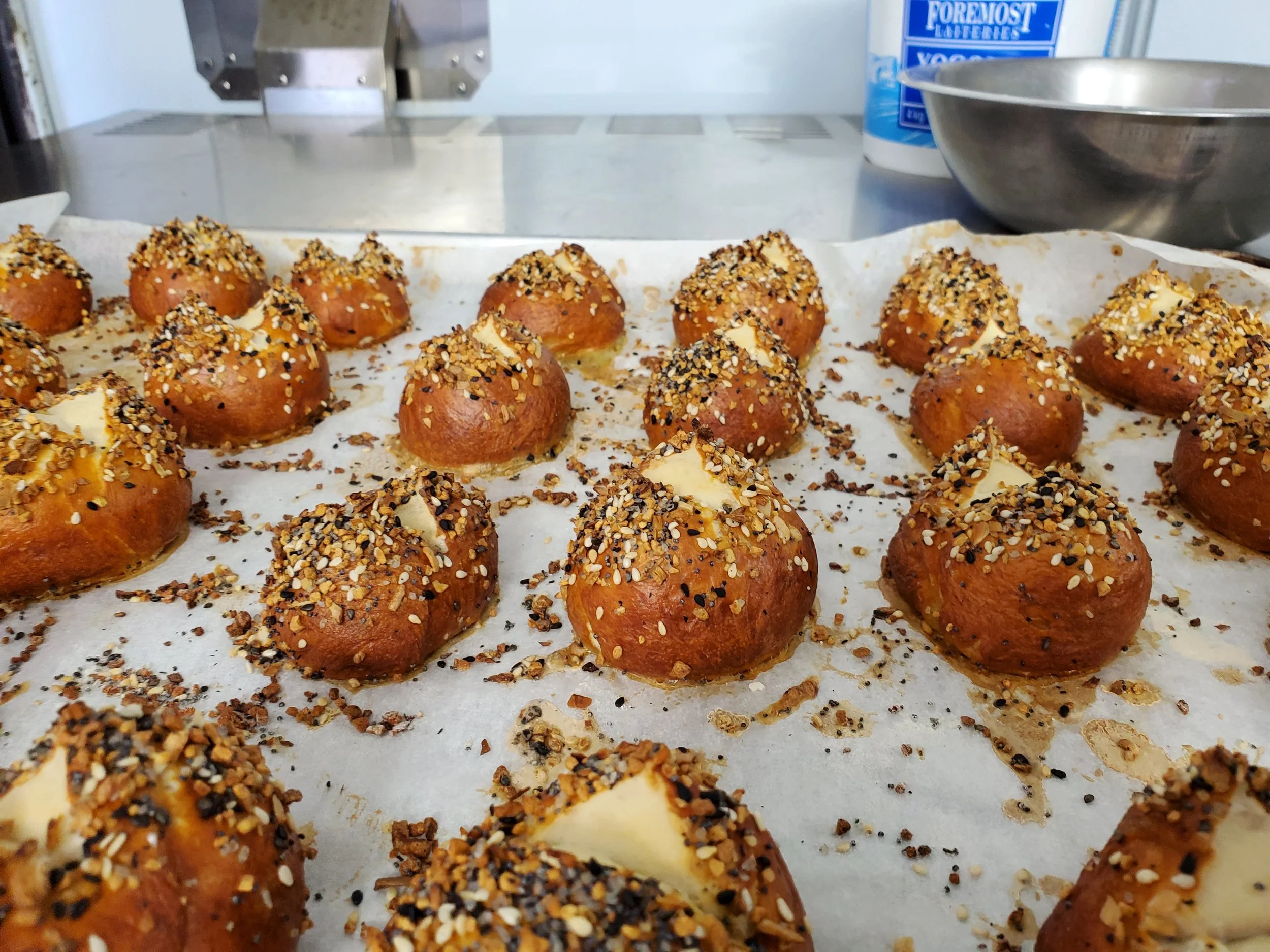 Tray of freshly baked bread rolls topped with chopped nuts and seeds, some with melted cheese underneath, on a baking sheet lined with parchment paper in a kitchen.