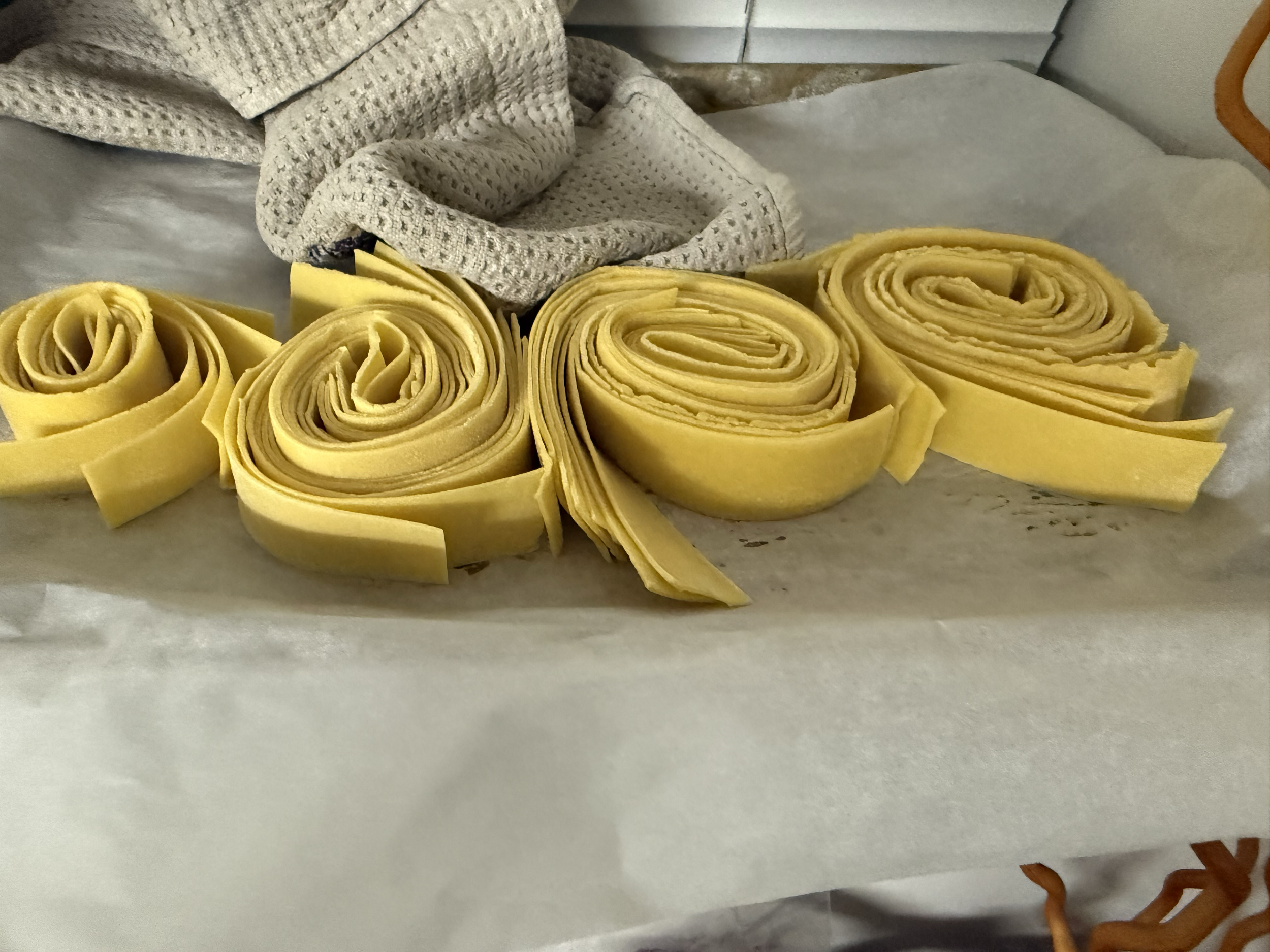 Several yellow pasta sheets rolled into spirals are placed on parchment paper with a textured cloth and a metal surface in the background.