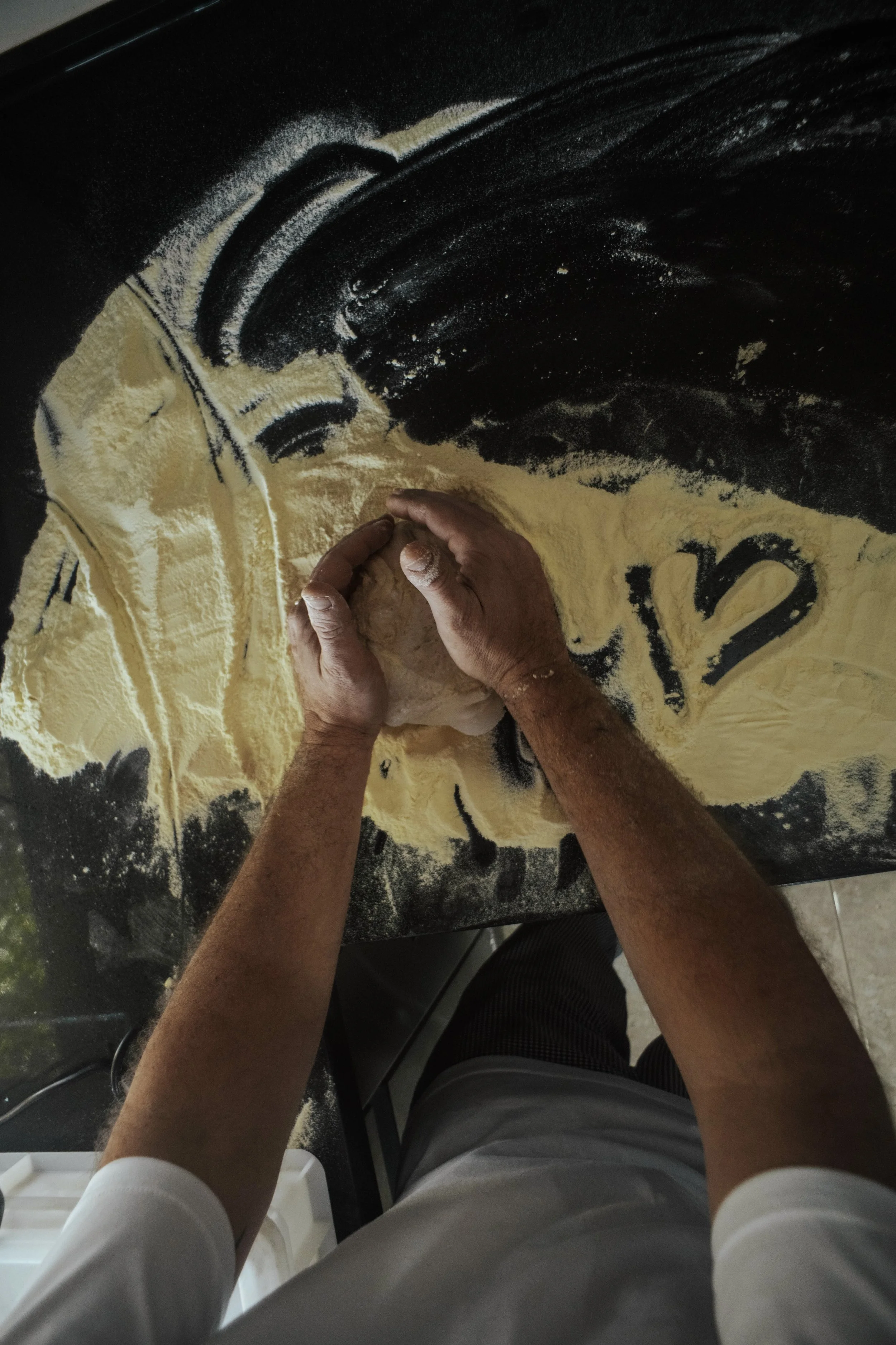 The Pizzaiolo is rolling out dough on a black countertop sprinkled with flour. The photograph is taken from a top-down perspective, showing the person's arms and hands actively shaping the dough. The Pizzaiolo has drawn a heart in the flour