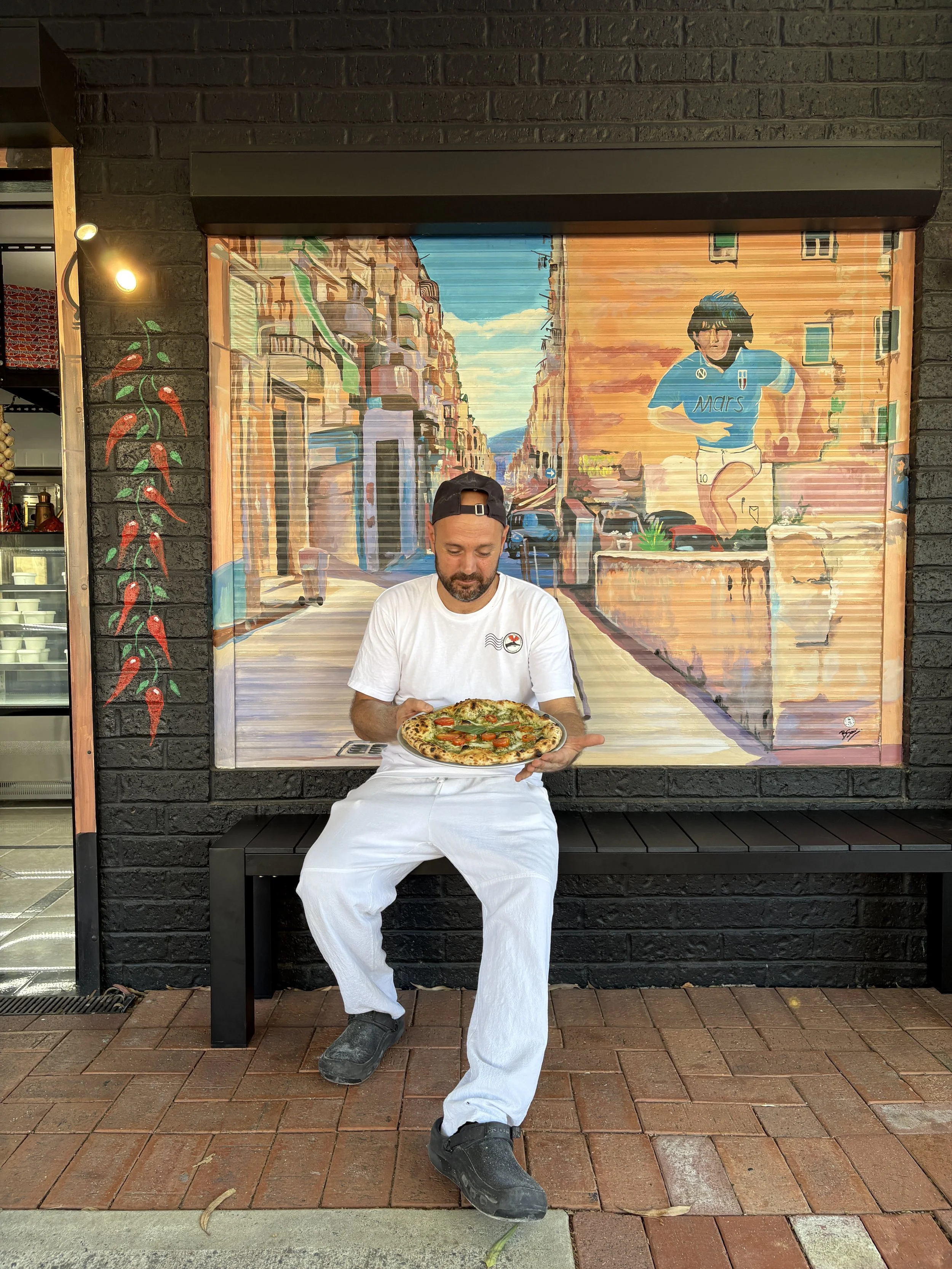 A pizzaiolo sitting outside his pizzeria admiring his pizza, next to a handprinted mural of Diego Maradona and Napoli's historical streets