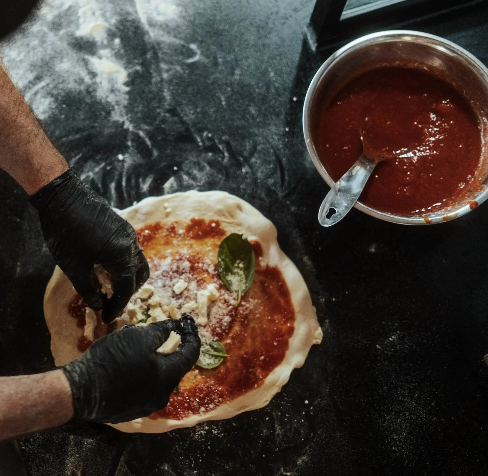 A Pizzaiolo making a pizza by adding Mozzarella and San Marzano sauce to dough, with tomato sauce in a bowl nearby, on a black countertop.