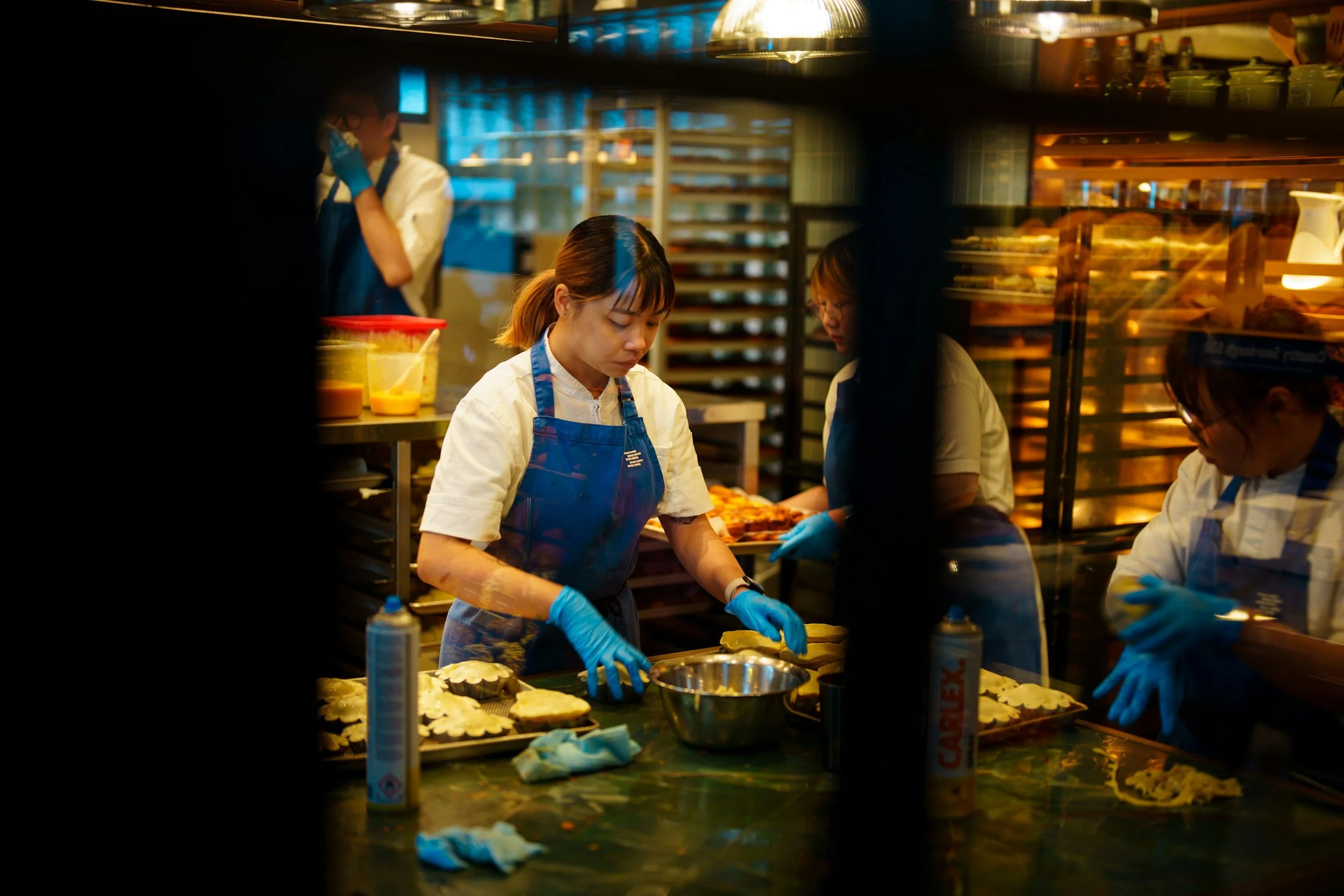 Behind a glass window, there are several people working in a bakery or pastry shop. They are wearing white coats and blue gloves, preparing baked goods on a dark green countertop. In the background, shelves are filled with baked items, and there is a person standing on the left side. The scene is lit with warm yellow lighting.