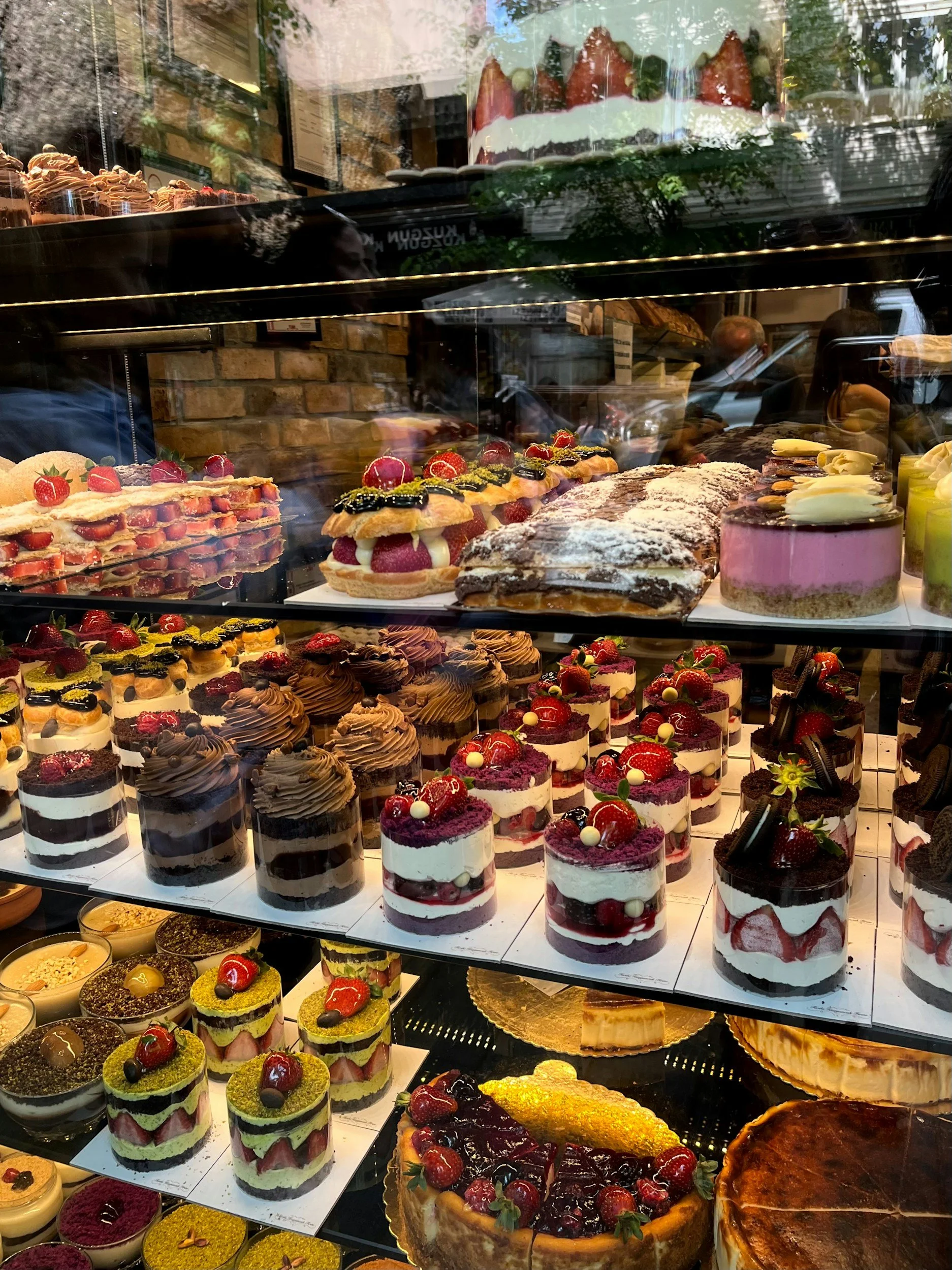 Assorted desserts in a bakery display case, including layered cakes with strawberries and cream, fruit tarts, chocolate mousse, and cheesecakes.
