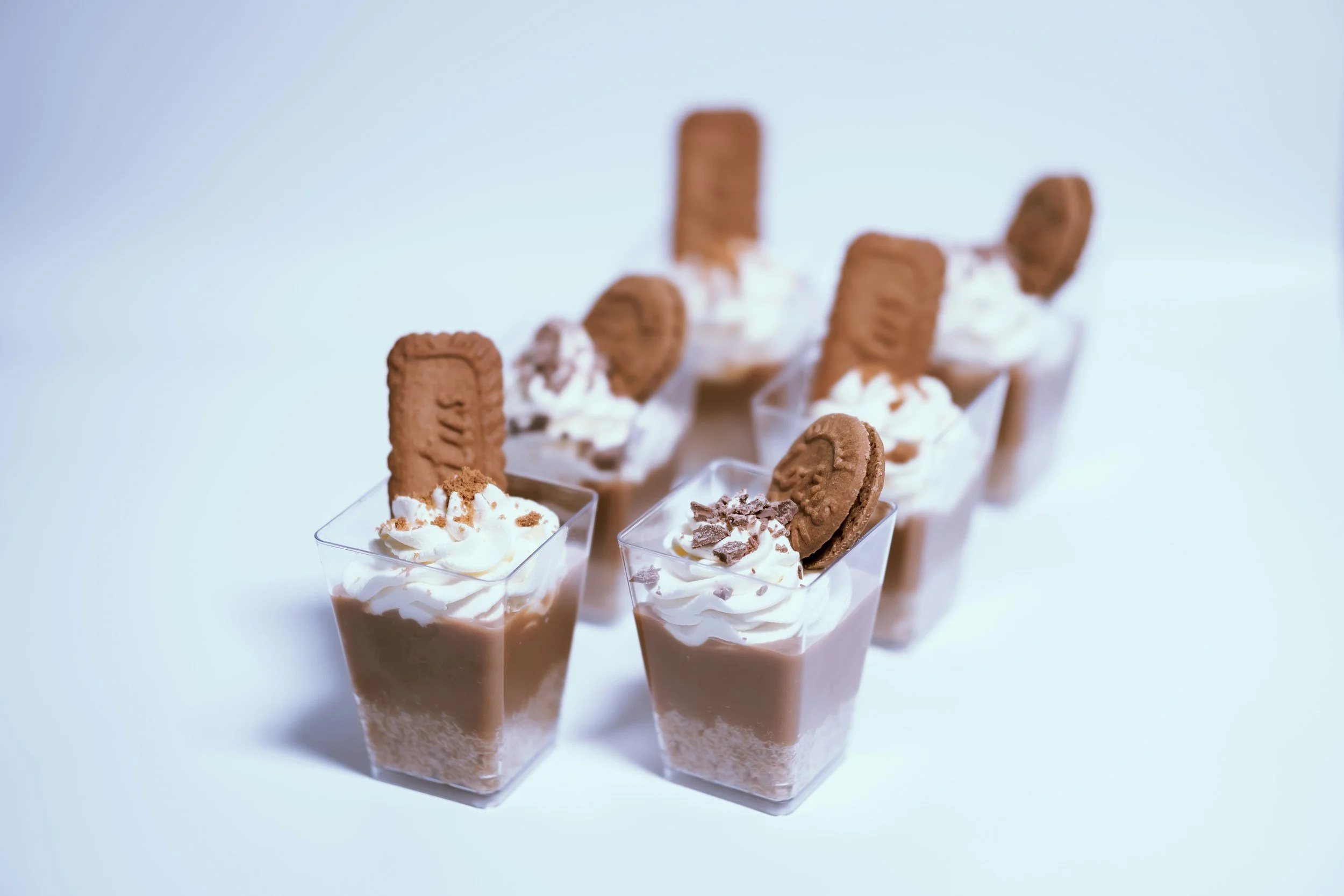 Six small clear plastic cups filled with chocolate mousse topped with whipped cream, chocolate shavings, and cookies, arranged in a row against a plain white background.