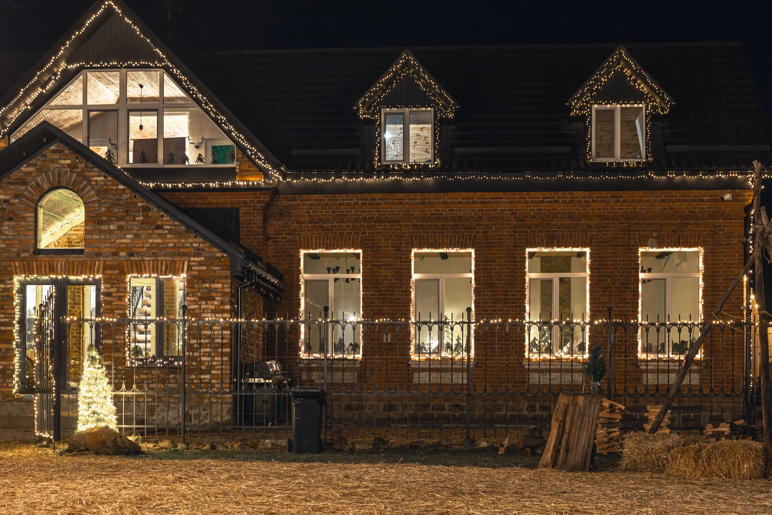 A house decorated with Christmas lights at night, with a brick exterior, multiple windows, and a steep roof with dormer windows illuminated by string lights.
