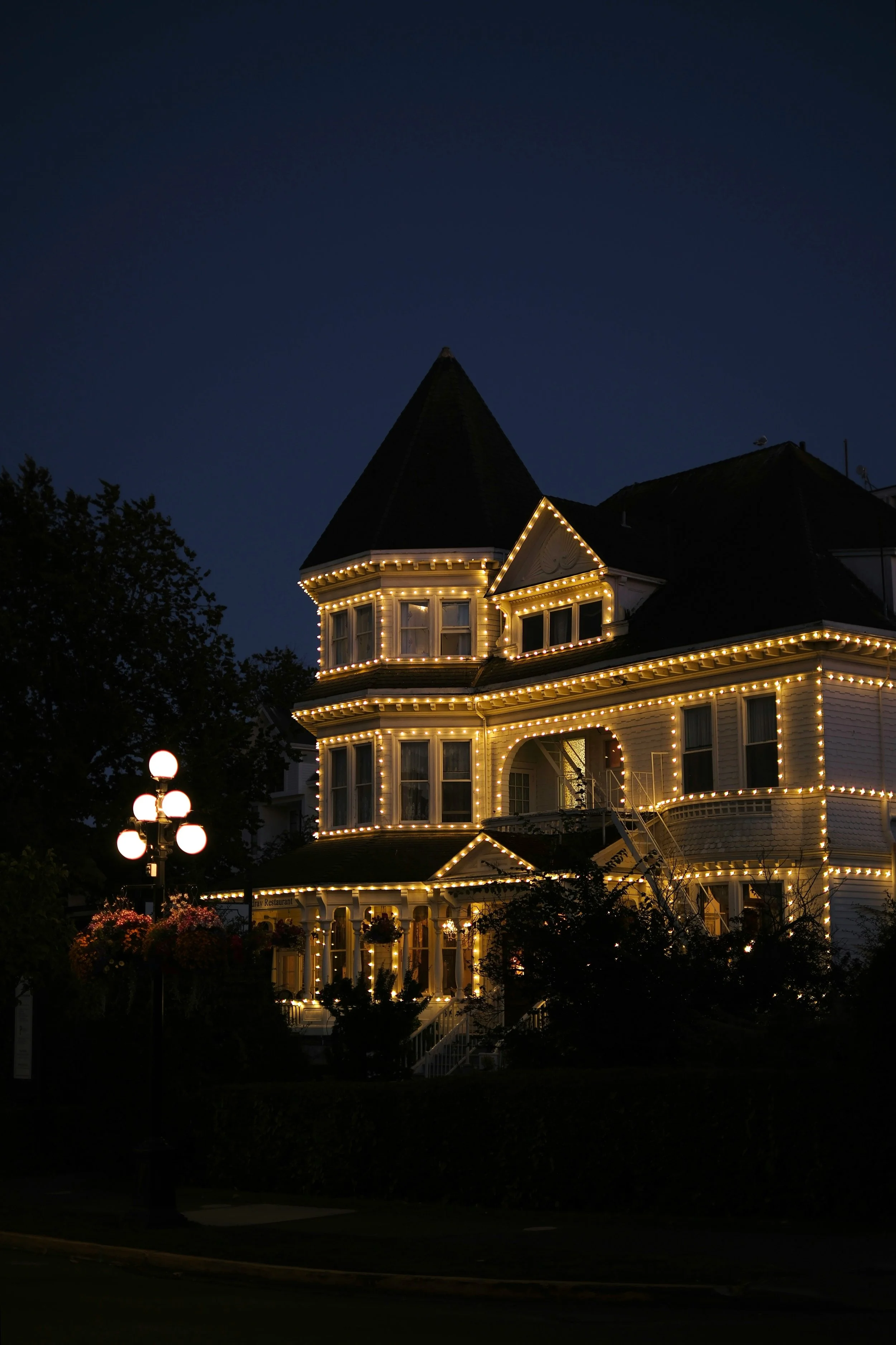 A Victorian-style house at night decorated with strings of warm white lights outlining its roof, windows, and porch. There are trees and a street lamp with glowing bulbs nearby.