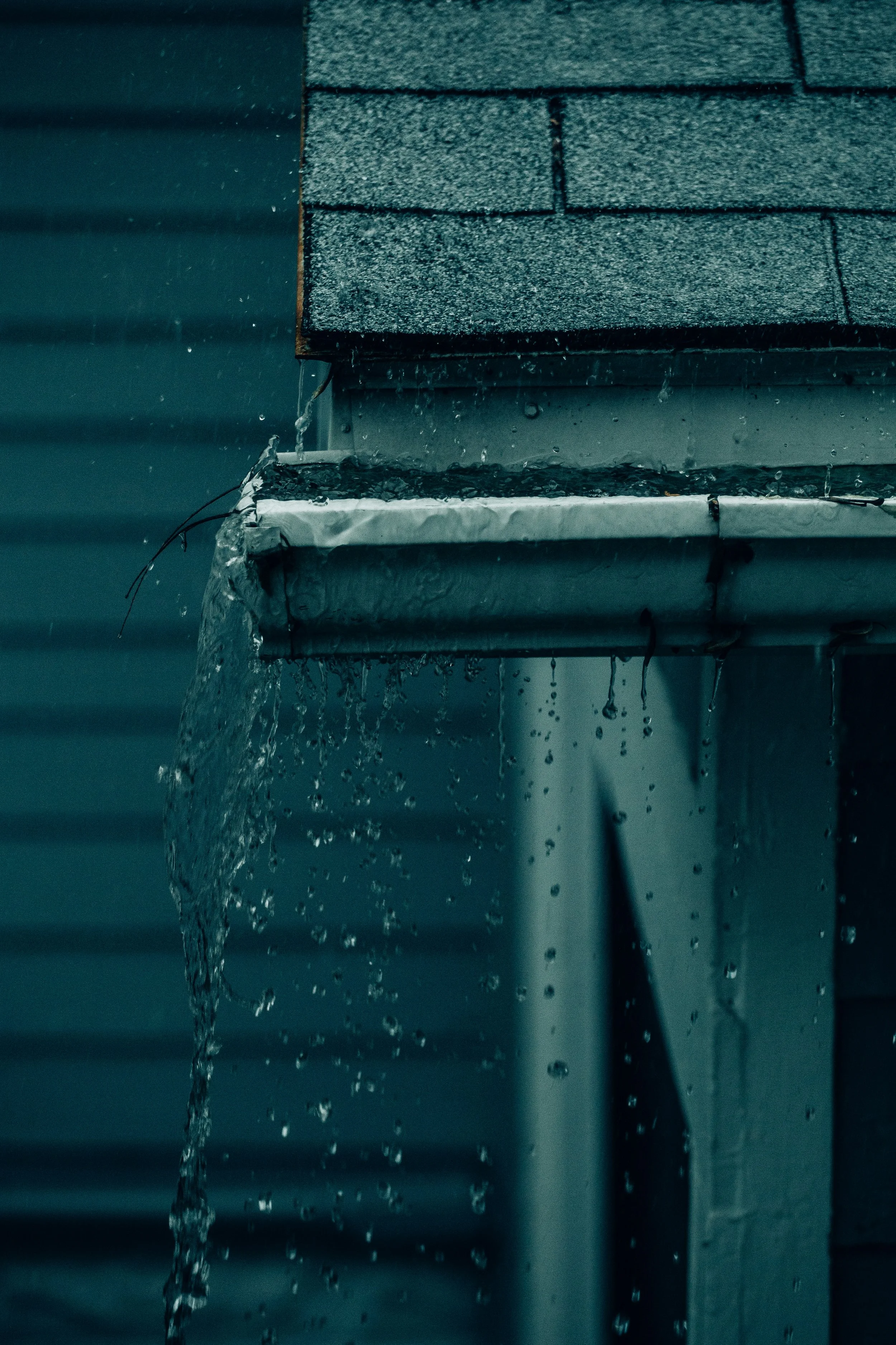 Water dripping from the edge of a roof with shingles, against a background of a rain-soaked house wall.