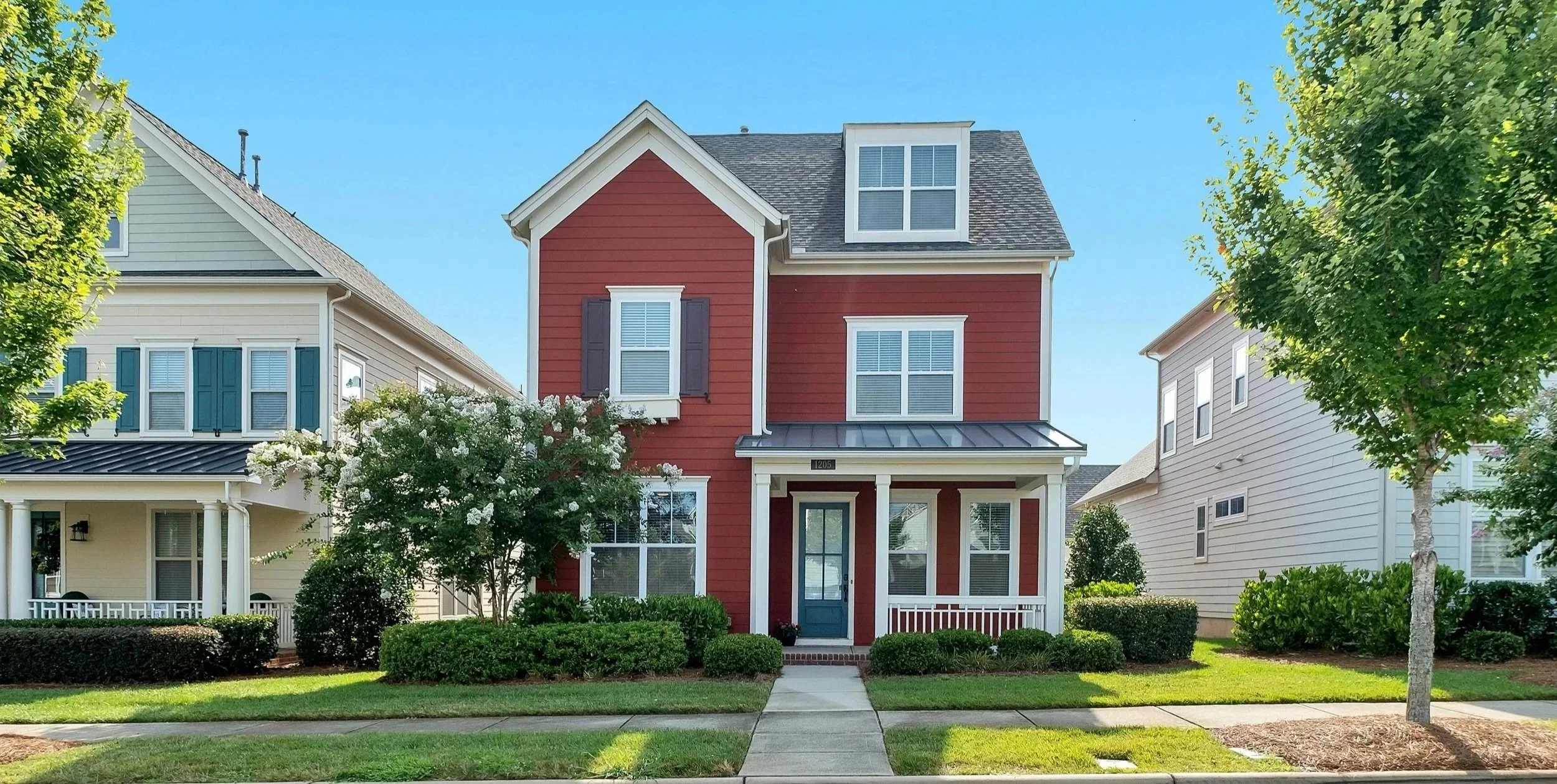 A red three-story house with white trim, black shutters, and a small front porch, surrounded by green trees and bushes, under a clear blue sky.
