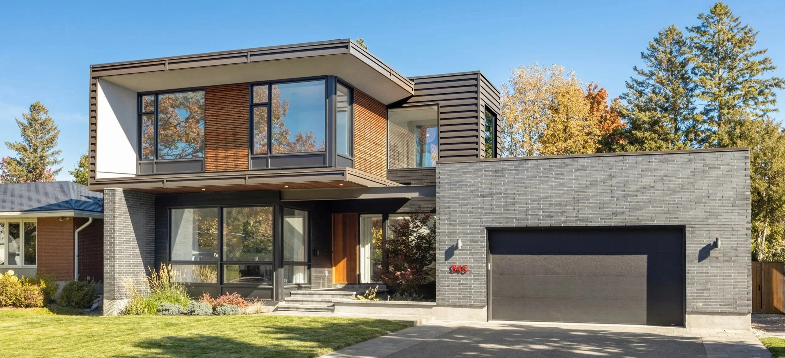 Modern two-story house with black brick and wood accents, large windows, and a gray garage door, situated in a landscaped yard with trees in the background.