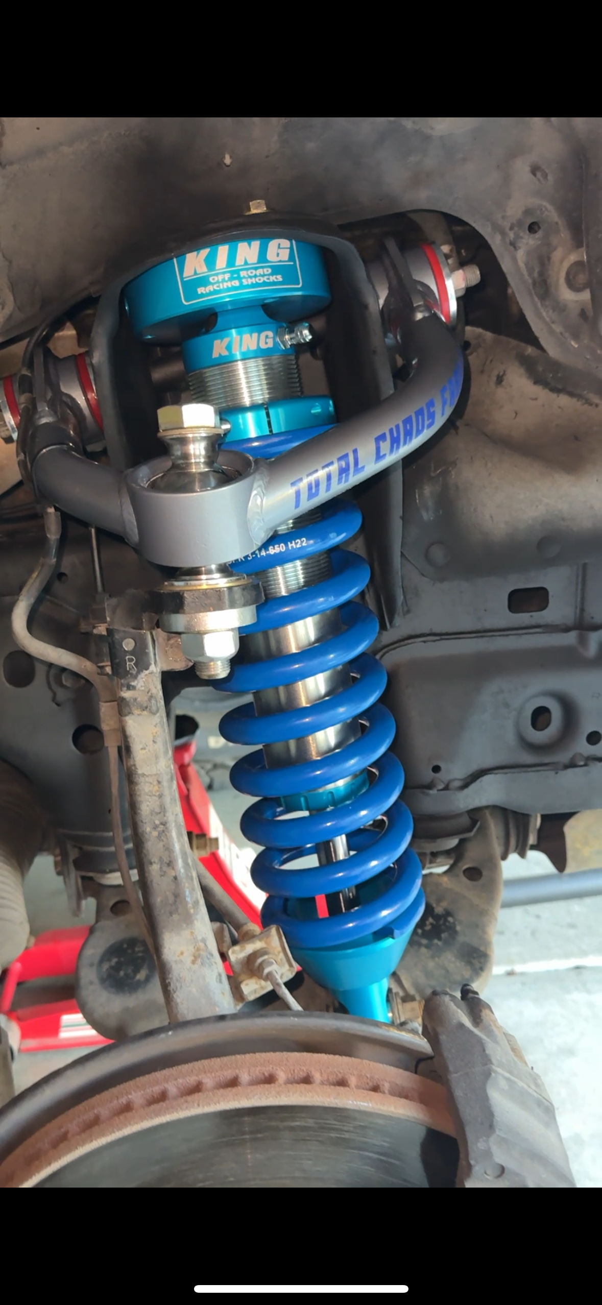 Close-up of a blue coilover suspension system on a vehicle, with visible shock absorber and parts labeled 'KING' and 'TOTAL CHAOS FAB.