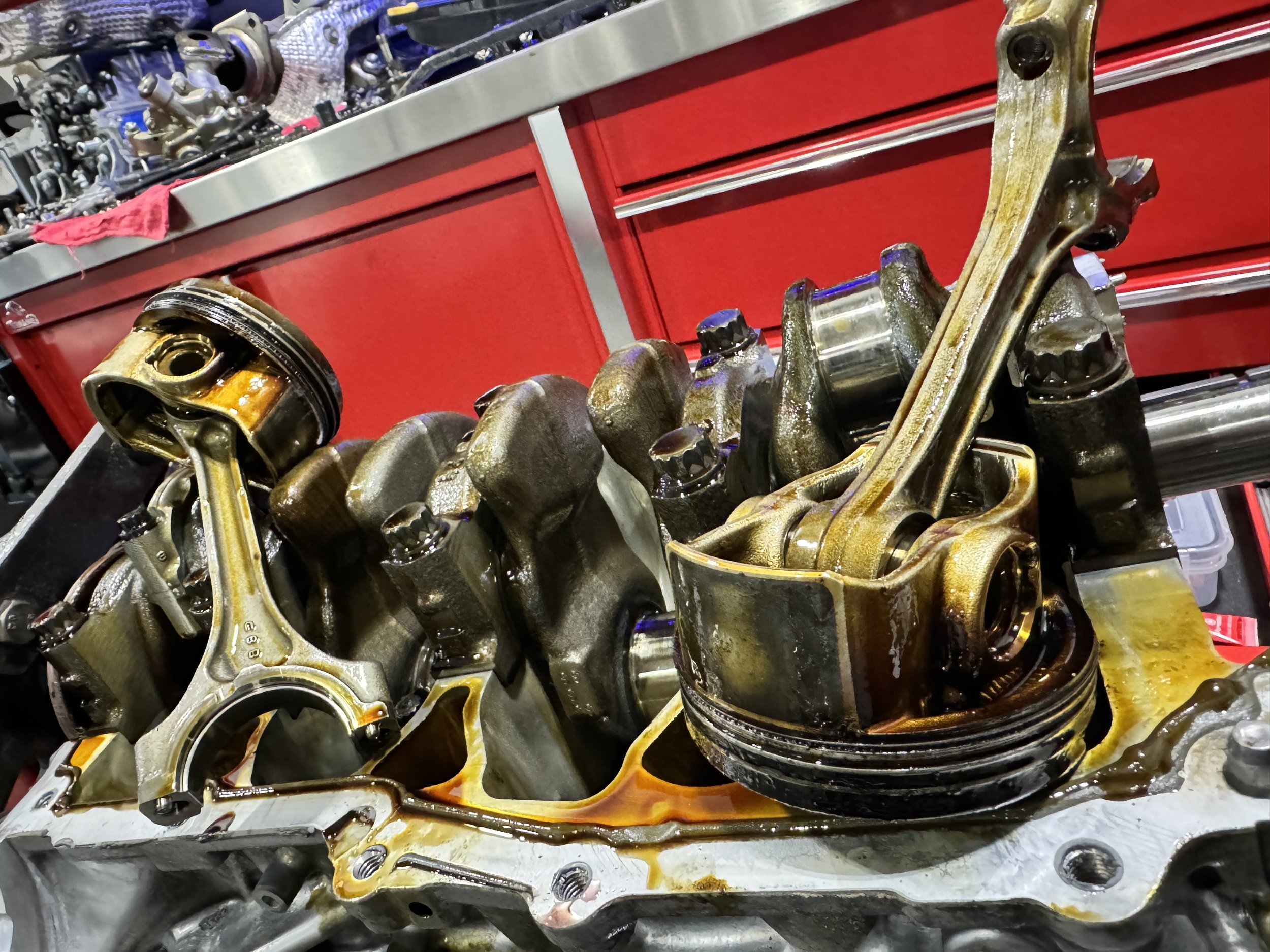 Disassembled engine parts with oil and grease, placed on a workbench with red tool storage cabinets in the background.