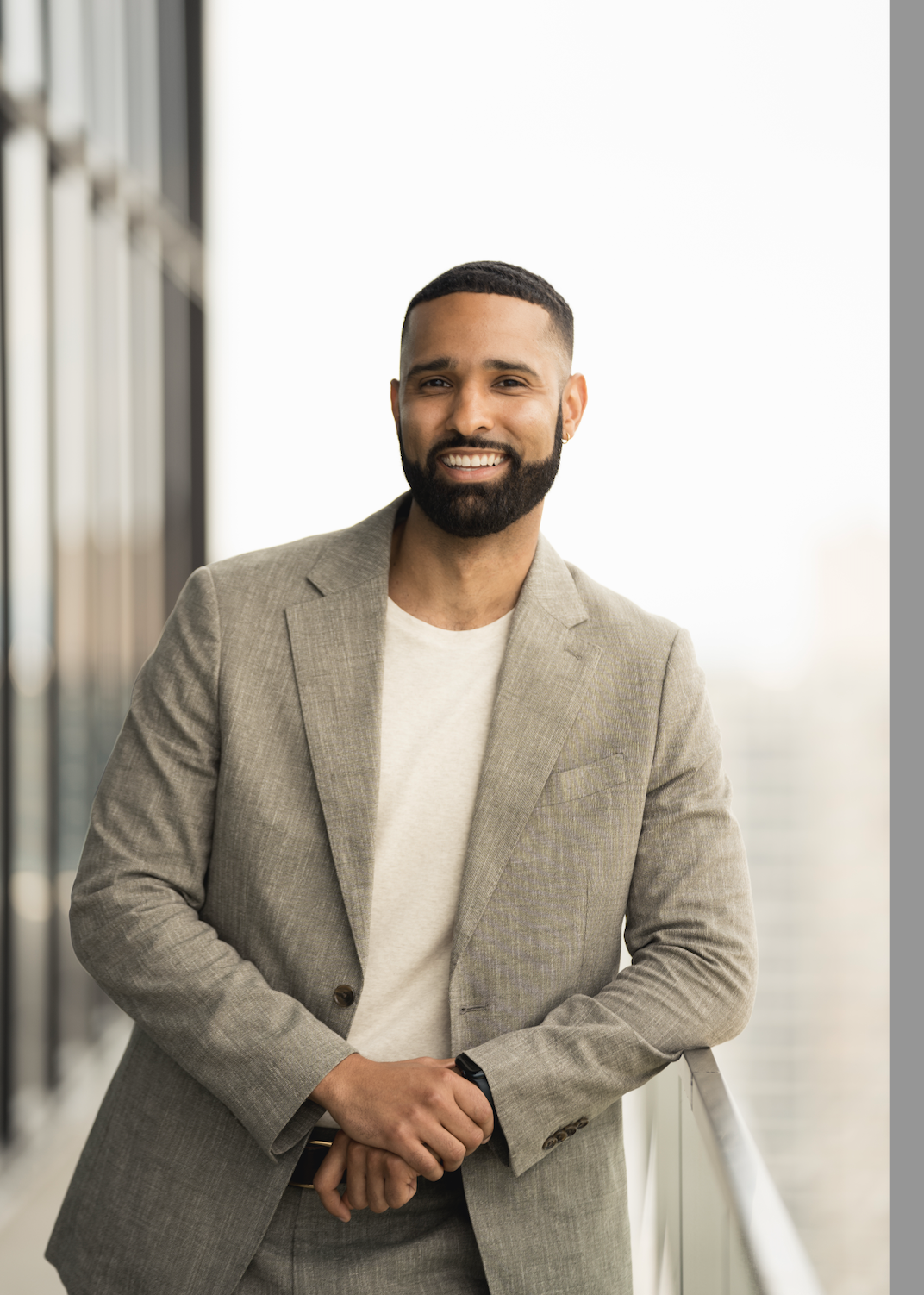 Smiling man with beard and short hair wearing a beige blazer and white t-shirt, standing on a balcony with a city skyline background in New York City.