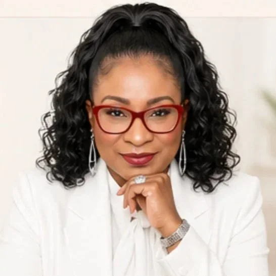 A woman with curly black hair wearing red glasses, jewelry, and a white outfit, sitting with her chin resting on her hand and smiling confidently.
