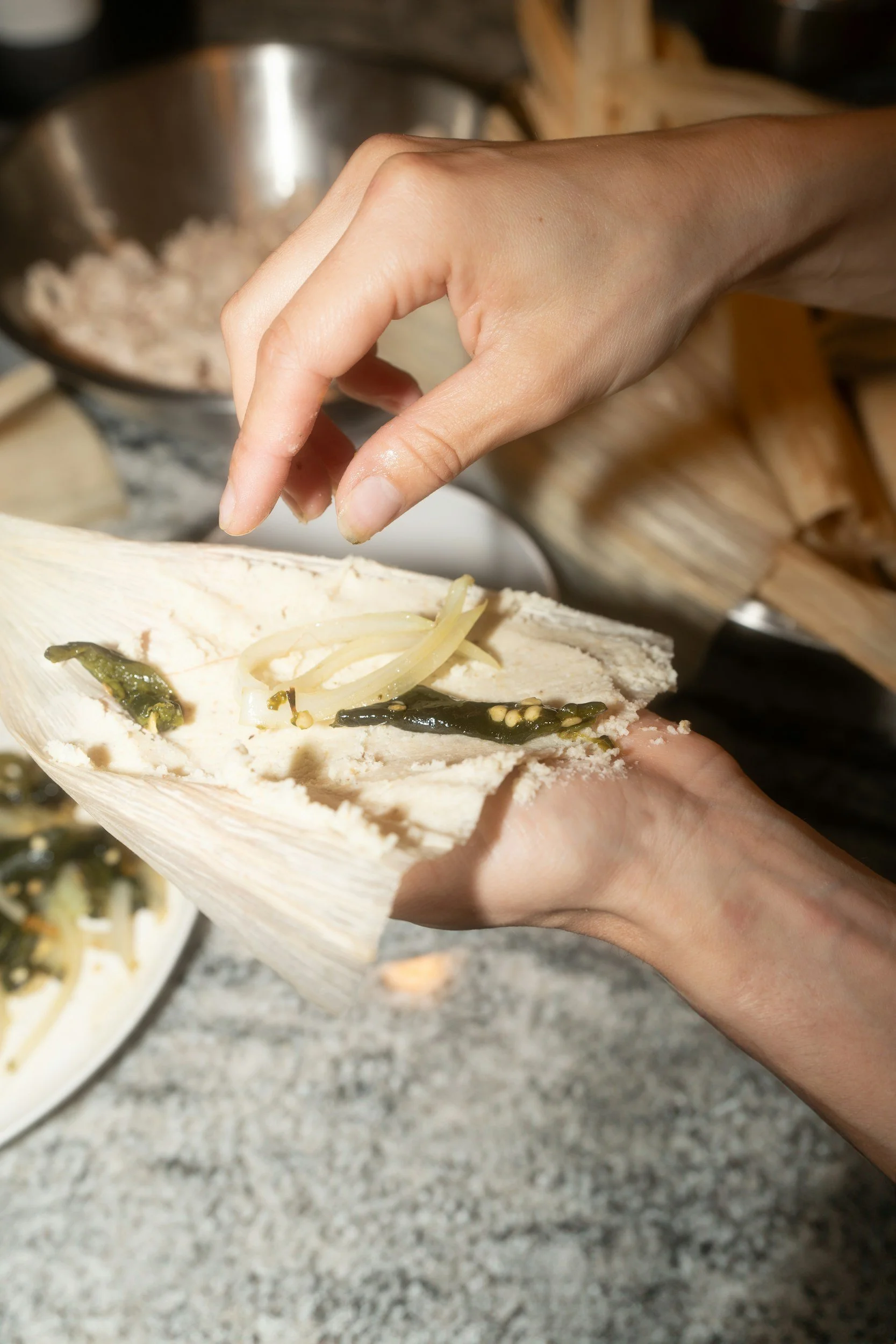 Person spreading corn masa dough on a corn husk to make Mexican tamales with a filling of chili peppers and onions.