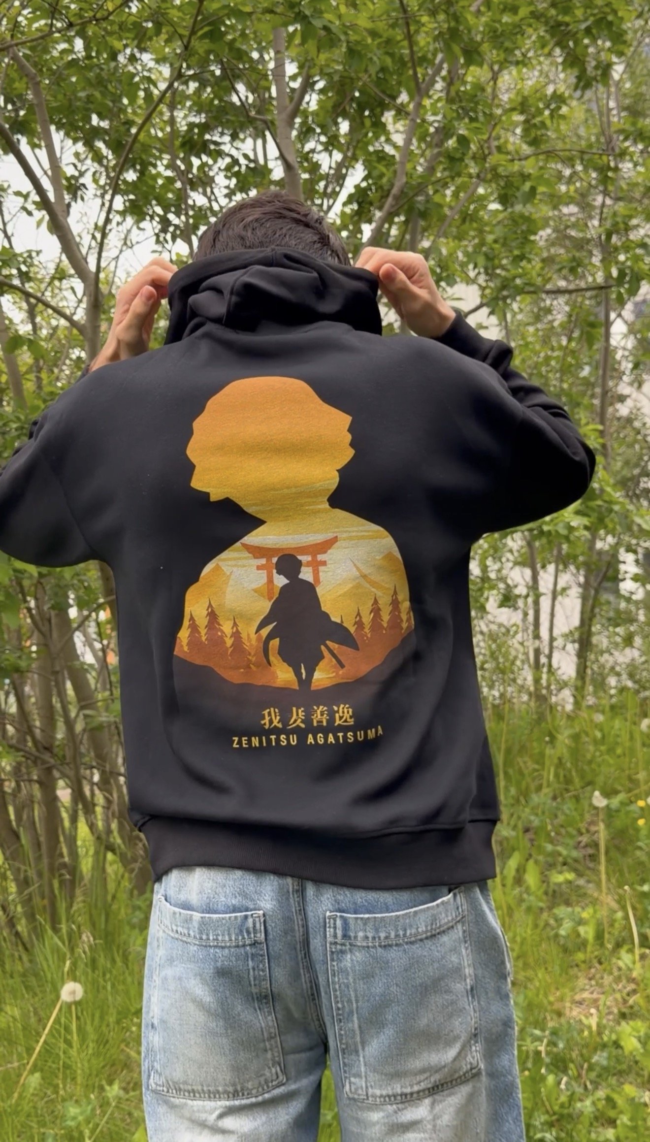 Person seen from behind, adjusting the collar of a black hoodie with a graphic design, in a green outdoor setting.