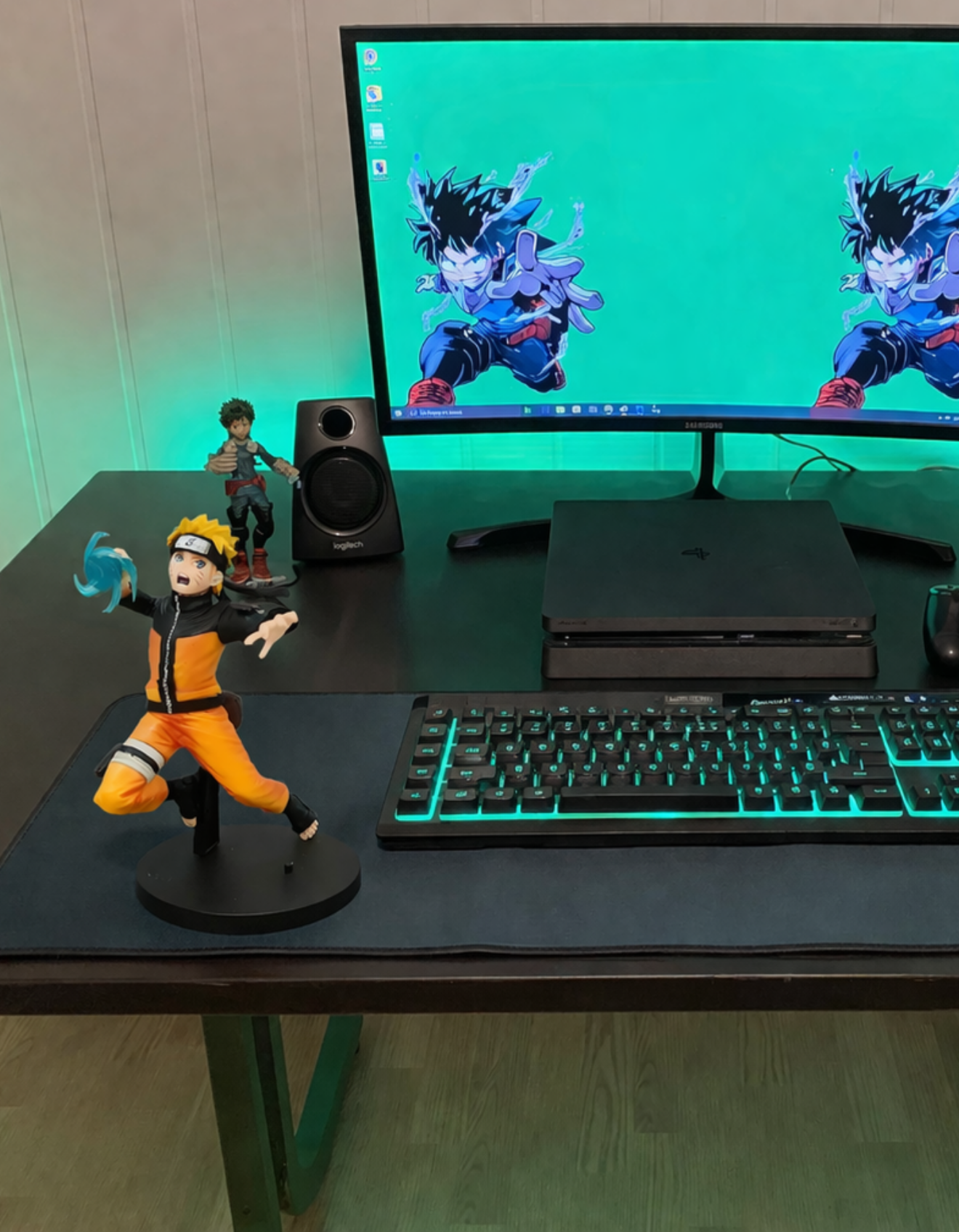 A computer desk setup with a monitor displaying a blue background with an animated character in a crouching pose, accompanied by figurines of characters from anime or manga, a keyboard with teal backlighting, a PlayStation console, a Logitech speaker, and a mouse.