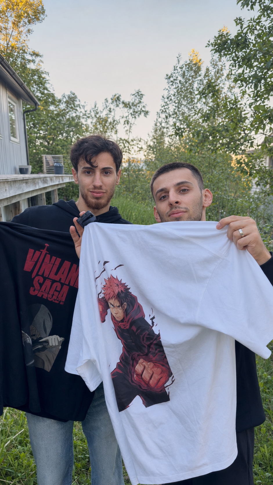 Two young men standing outdoors, each holding up a t-shirt with anime character graphics. The man on the left holds a black shirt featuring a character from 'Vigilante Saiyan,' and the man on the right holds a white shirt with a character with red hair in a fighting pose. They are in a garden or backyard setting with trees and a house in the background.