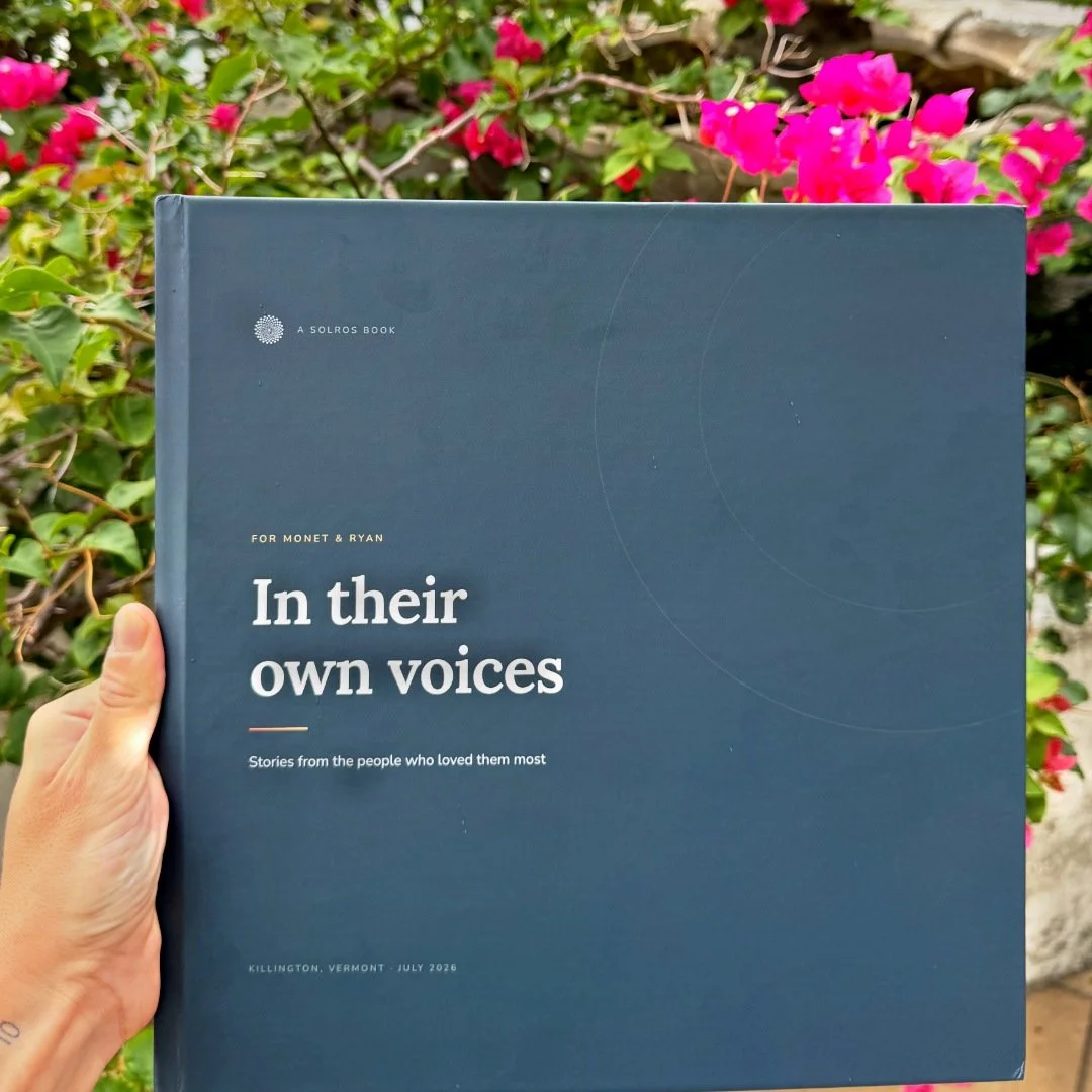 A person's hand holds a dark blue book titled 'In their own voices' in front of pink and red flowers and green foliage. This is the real product.