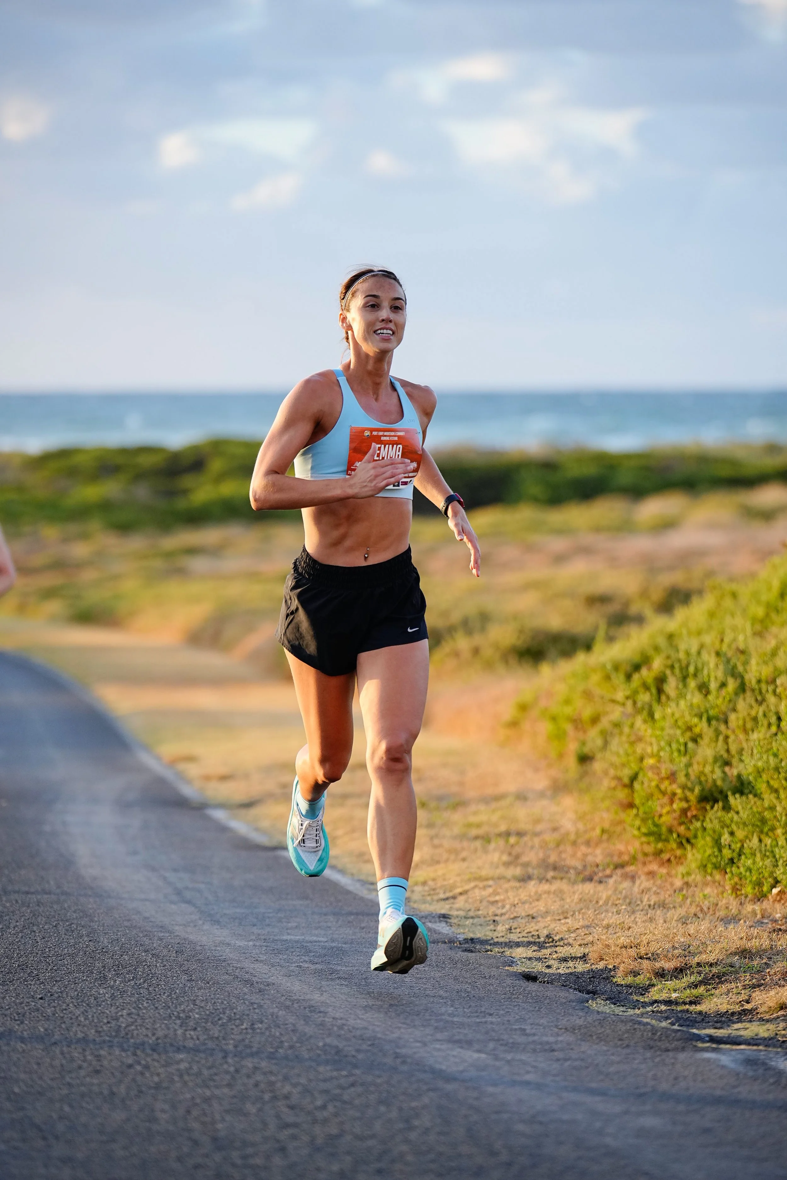 A female runner with a Port Fairy Marathon green race bib numbered 'EMMA' running along a paved trail near a grassy area with the Port Fairy ocean in the background, under a partly cloudy sky.