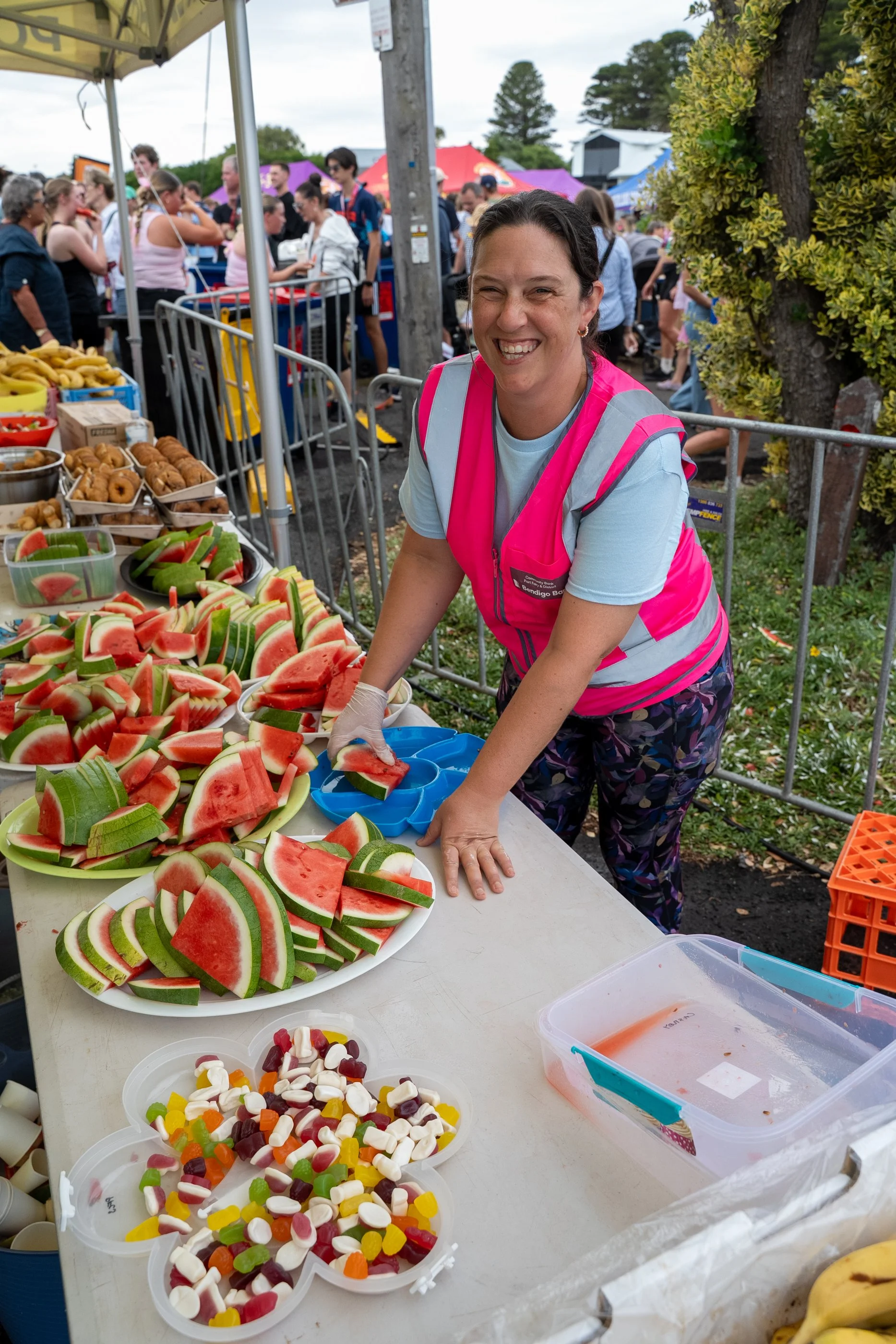 A smiling woman in a pink safety Port Fairy Marathon volunteer vest preparing watermelon slices at an outdoor event table, with various candies and pastries on the table, and a crowd of people and tents in the background.