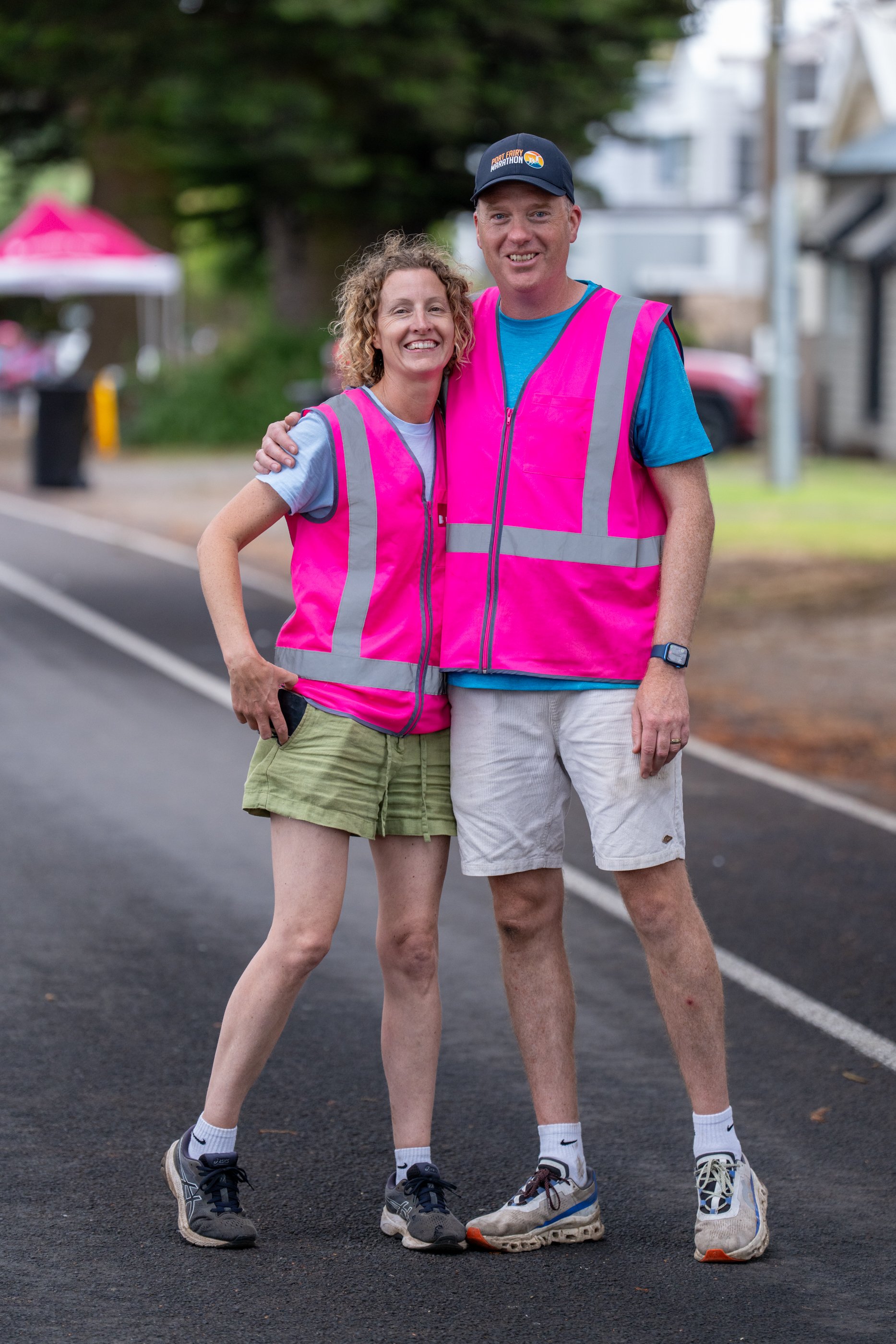 A smiling woman and man wearing bright volunteer Port Fairy Marathon vests, standing on a paved road outdoors in Port Fairy.