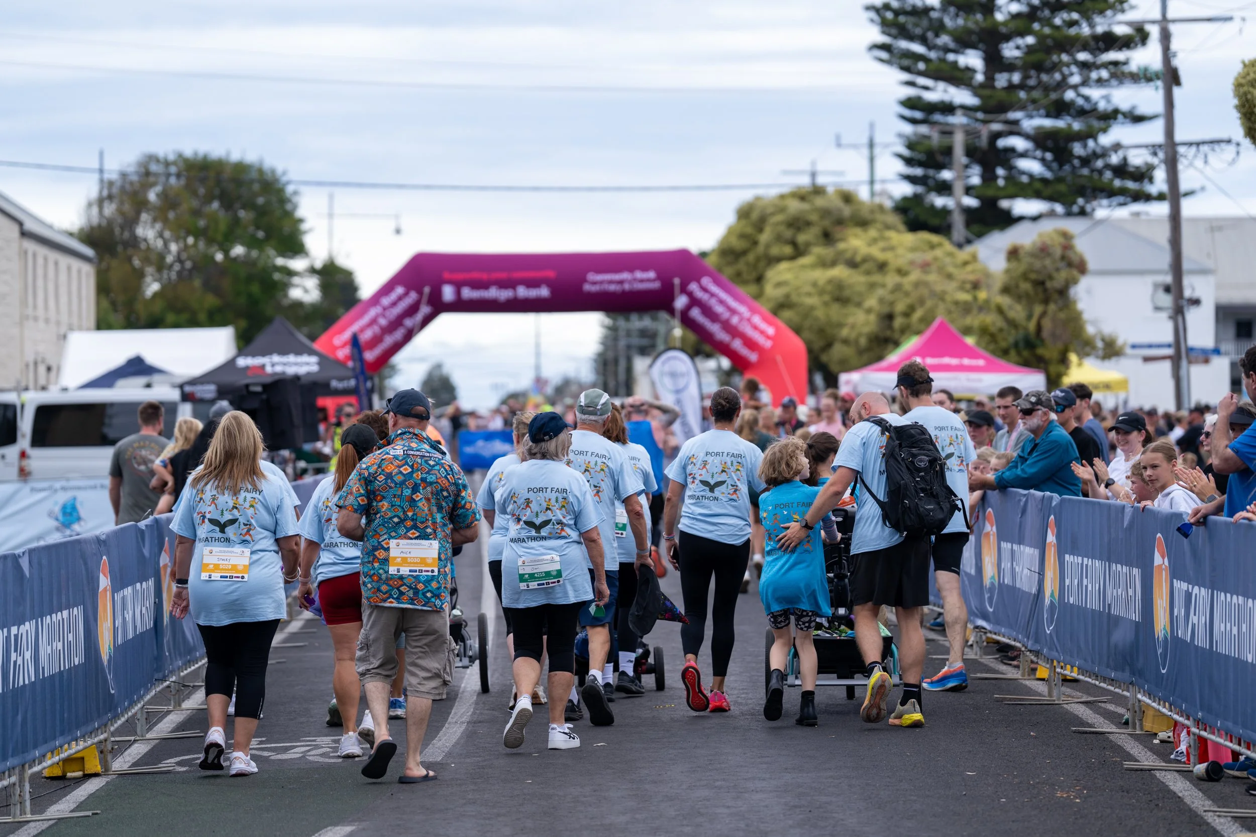 People participating in the Port Fairy Marathon event, walking along a designated route lined with banners, with some wearing event T-shirts that say 'Port Fairy Marathon' and race bibs, under partly cloudy skies.