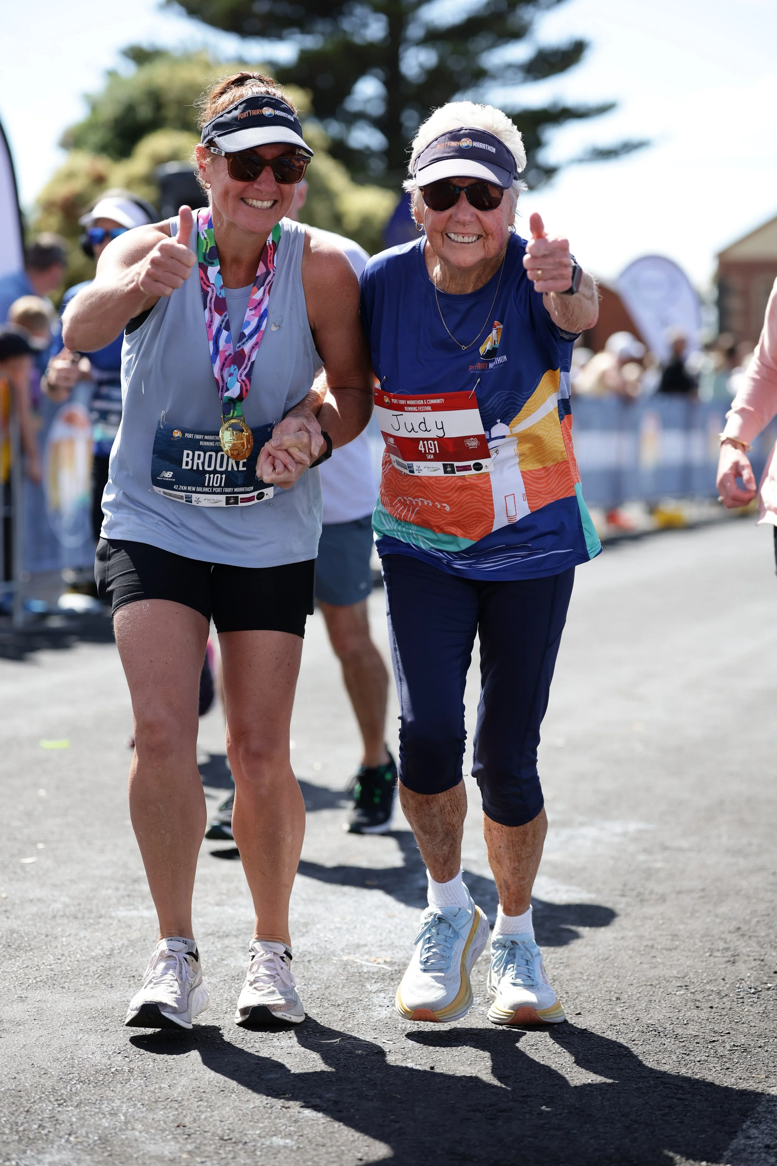 Two women running in the Port Fairy Marathon, smiling, giving a thumbs-up, wearing sunglasses, running attire, and Port Fairy Marathon race bibs, with trees and other runners in the background on a sunny day.