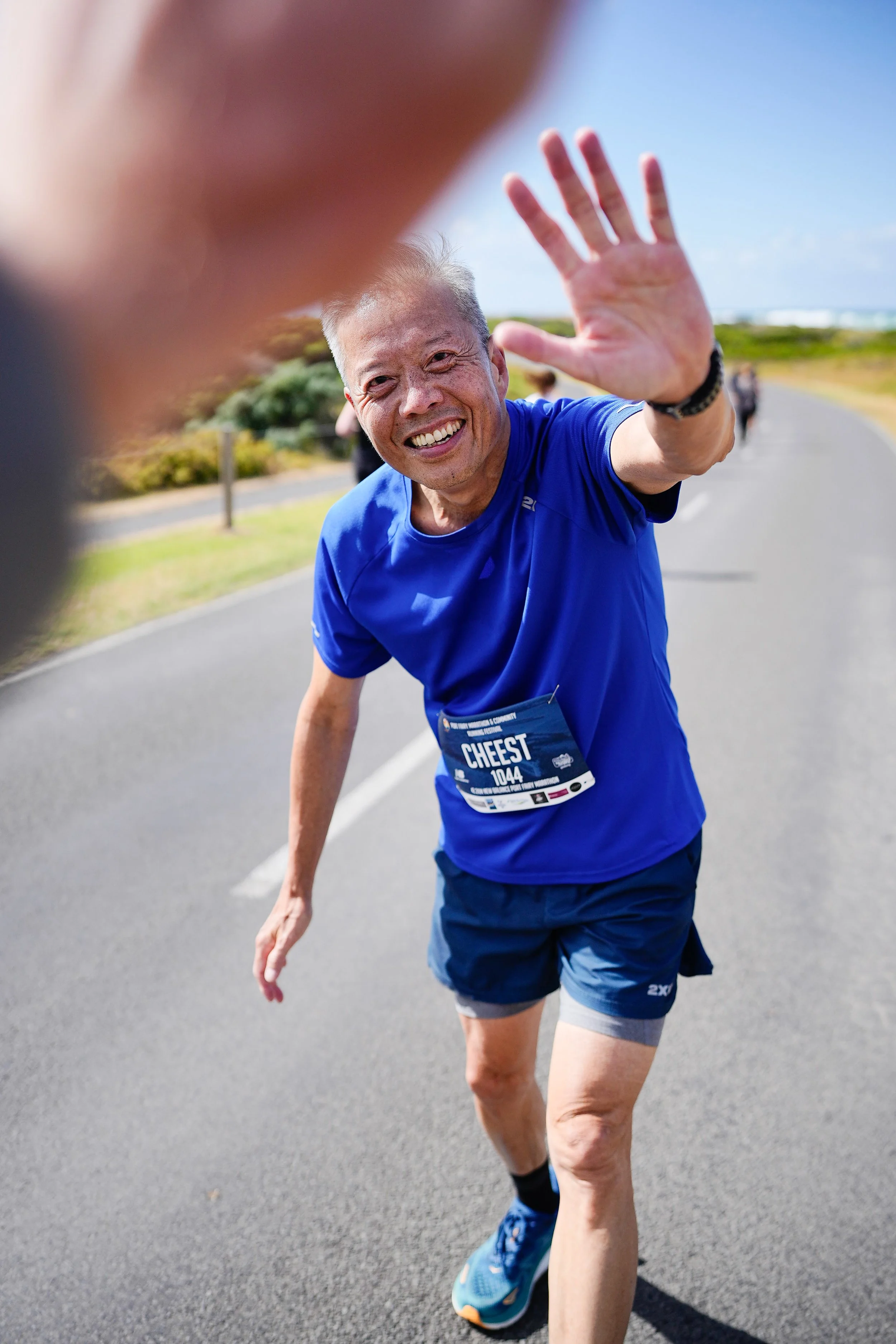 A man in a blue shirt and shorts running outdoors on a paved road at the Port Fairy Marathon, smiling and reaching towards the camera as he approaches it, with a blue sky and greenery in the background.