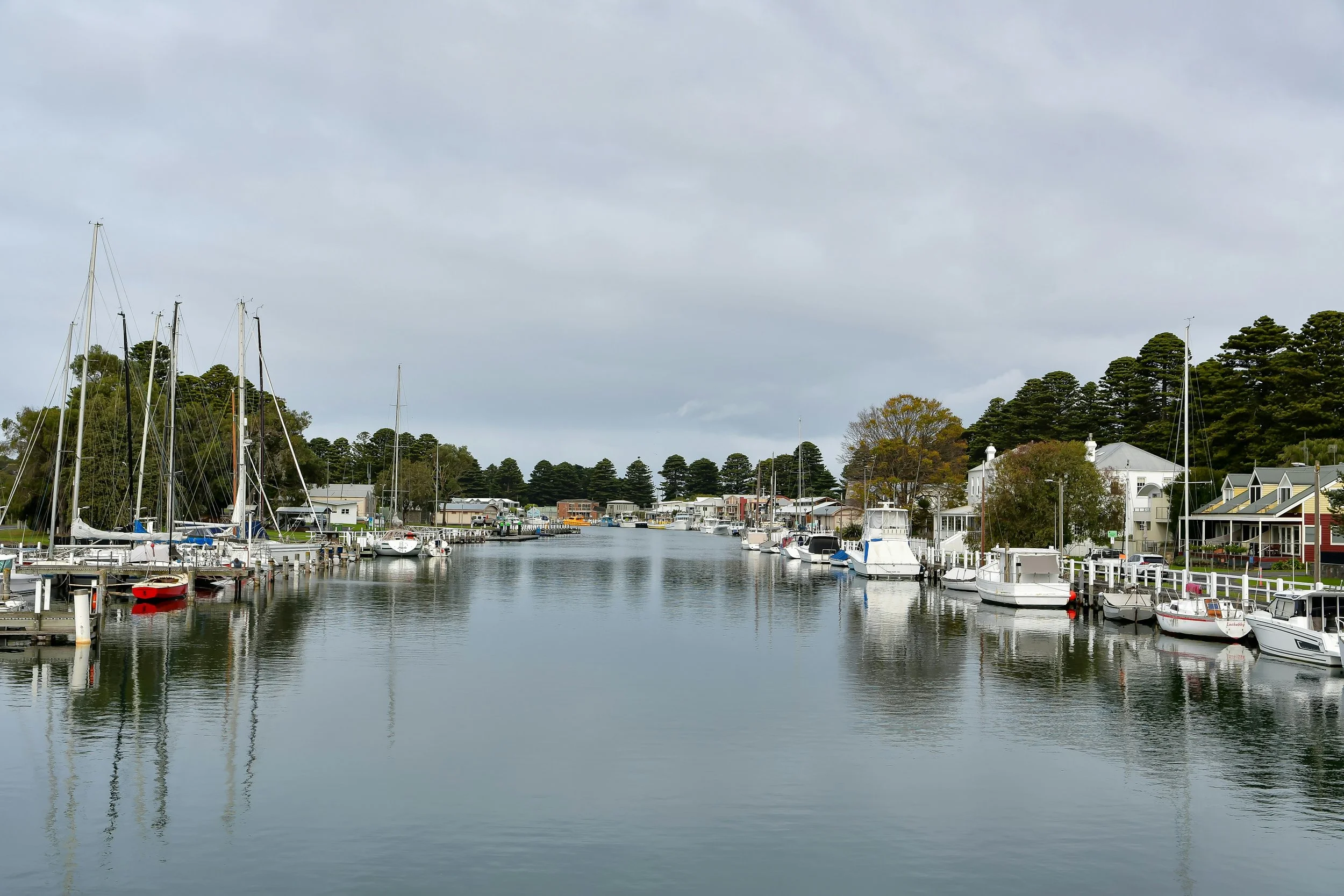 The Port Fairy marina with sailboats docked along the water, residential houses and trees in the background, and an overcast sky.