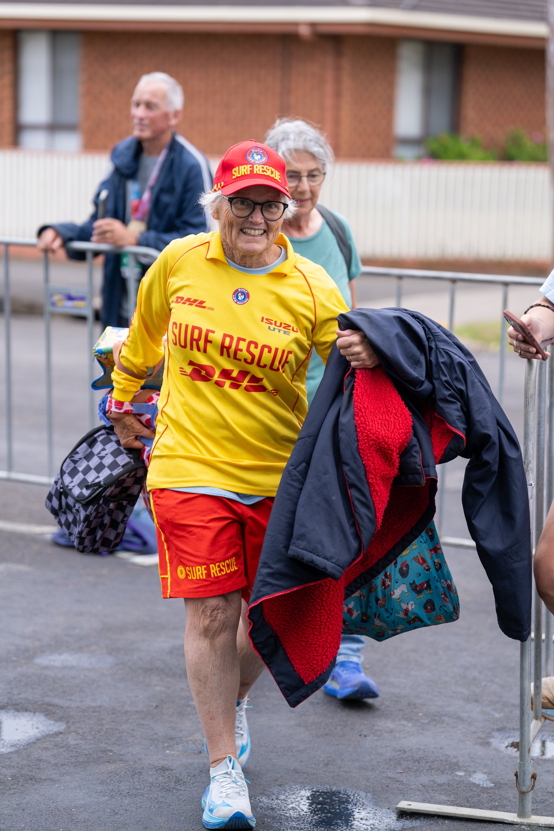 An woman surf life saving club volunteer dressed in a yellow surf rescue shirt and red shorts smiling and walking outdoors, holding a coat and a patterned bag on a gray pavement.