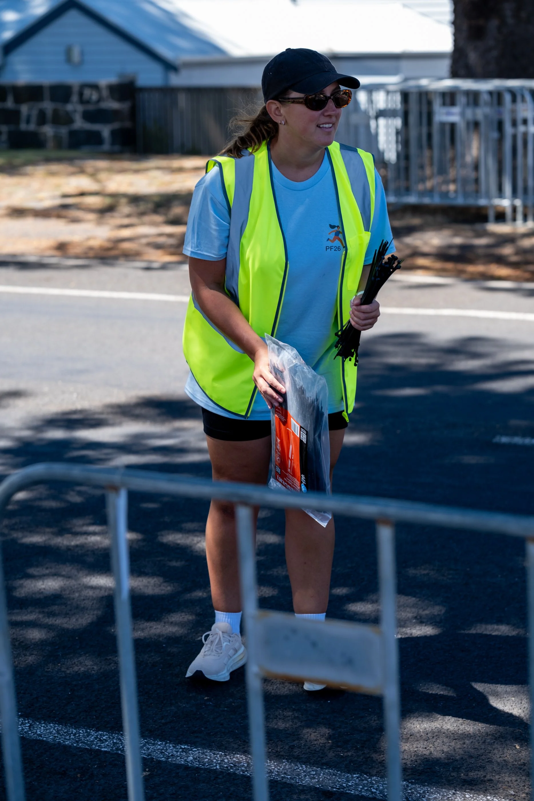 A woman wearing a yellow Port Fairy Marathon volunteer vest, sunglasses, a black cap, a light blue t-shirt, black shorts, and white sneakers, holding items and standing on a dark outdoor surface in front of a metal barricade.