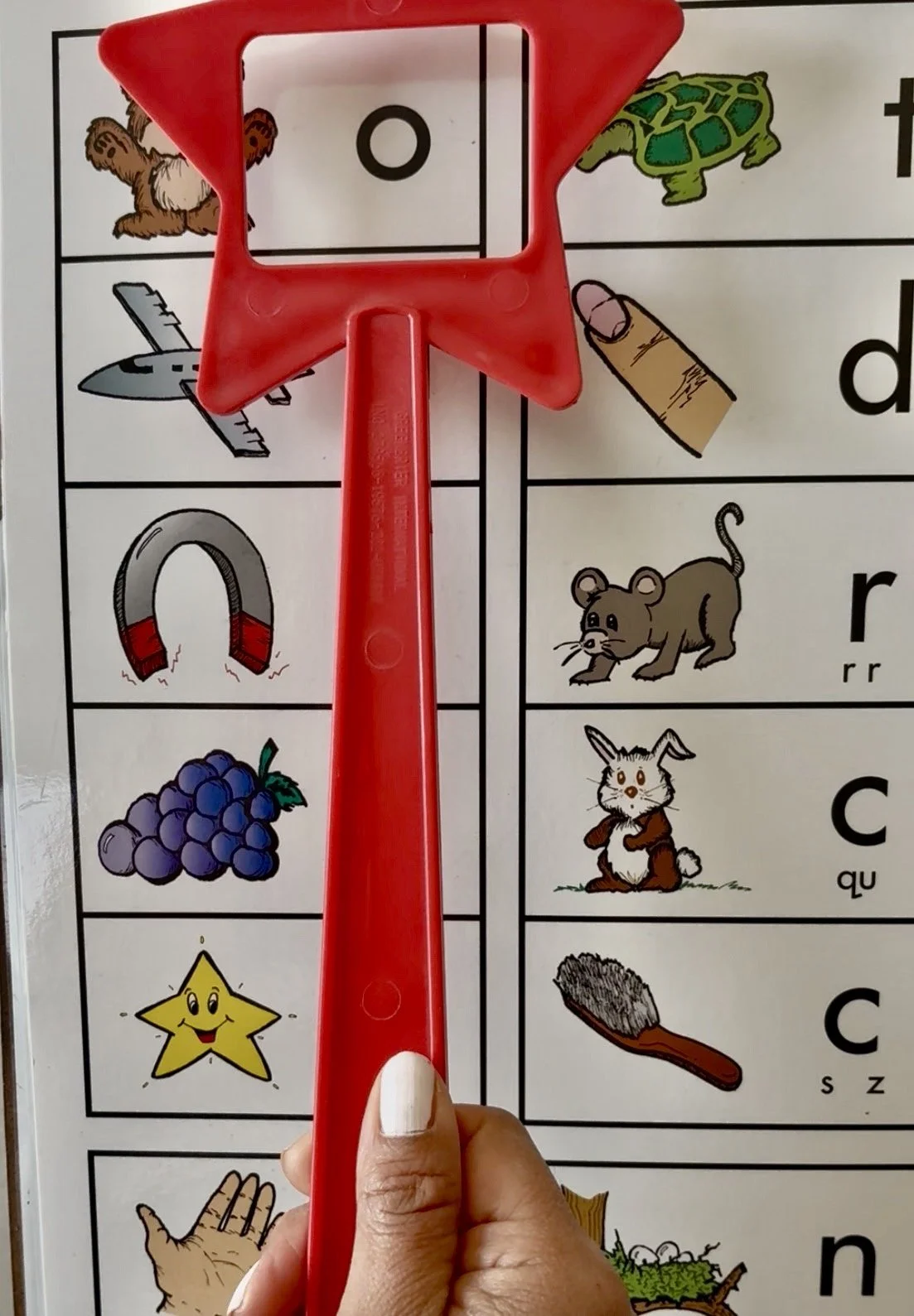 Close-up of a child's hand holding a red star-shaped puzzle piece over a classroom word chart with images of animals, objects, and vowels.
