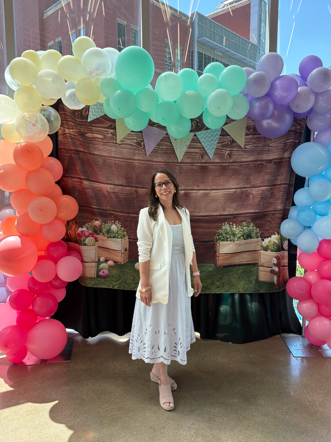 A woman in a white dress and blazer standing in front of a colorful balloon arch and a backdrop with potted flowers, outdoors under a clear blue sky.