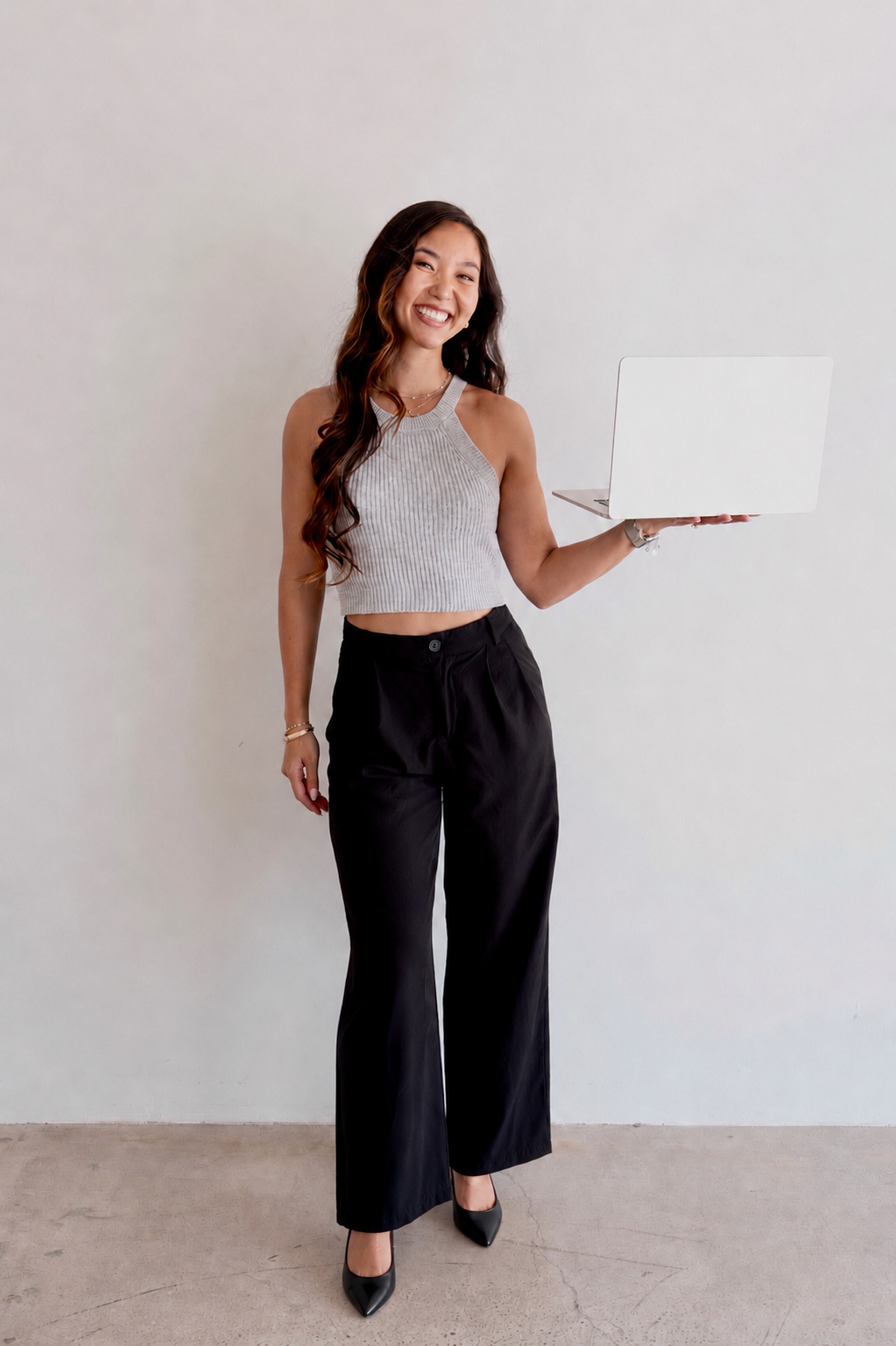 A woman standing on a plain light-colored wall, smiling, holding an open silver laptop in her right hand, dressed in a sleeveless gray crop top and black wide-leg pants with high heels.