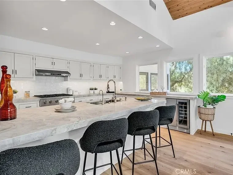 Modern kitchen with white cabinets, marble countertop island, black barstools, stainless steel stove, and large windows with a view of trees.