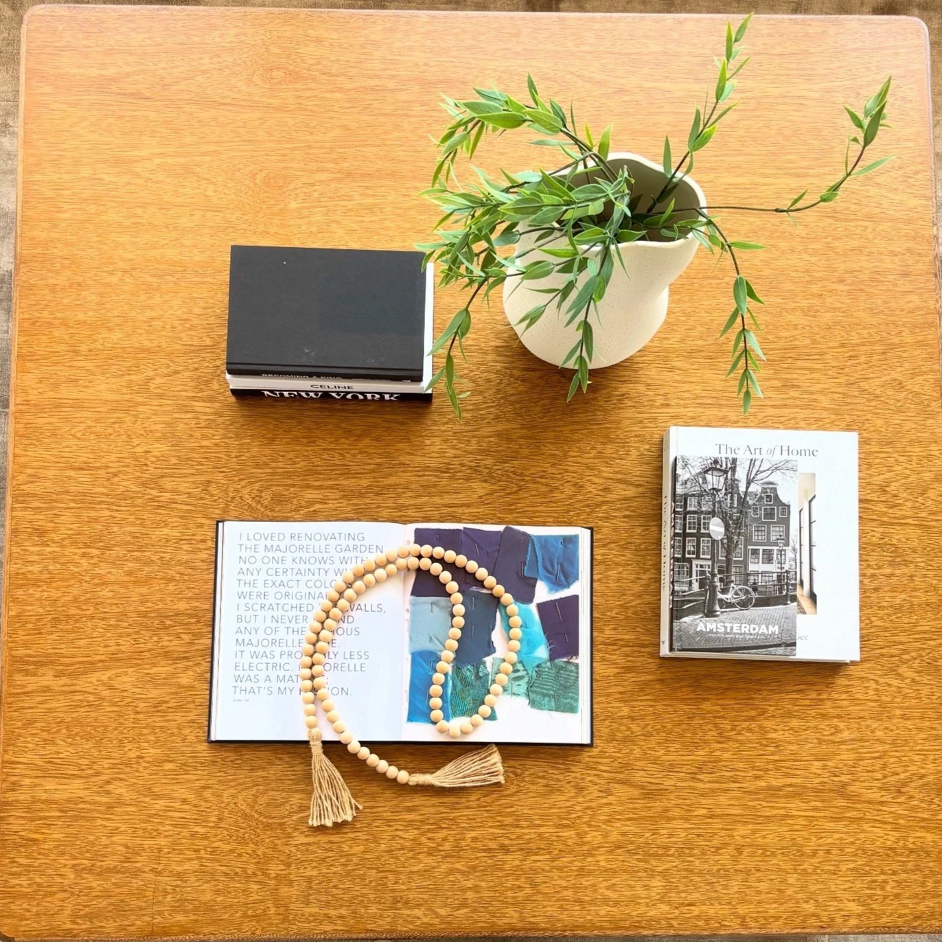 A wooden table with a potted plant, a stack of books, an open magazine with a beaded necklace, and a closed magazine.