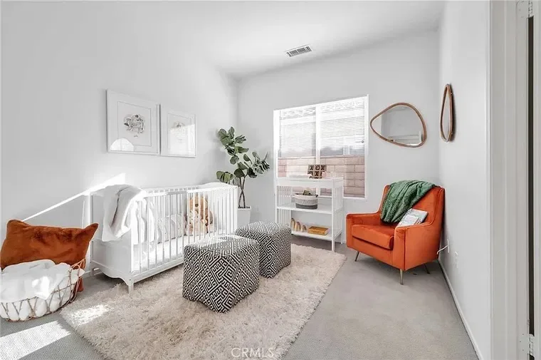 A nursery with white walls and a large window, featuring a white crib, potted plant, two patterned poufs, an orange armchair with a green throw, a white bookshelf, and framed artwork on the wall.