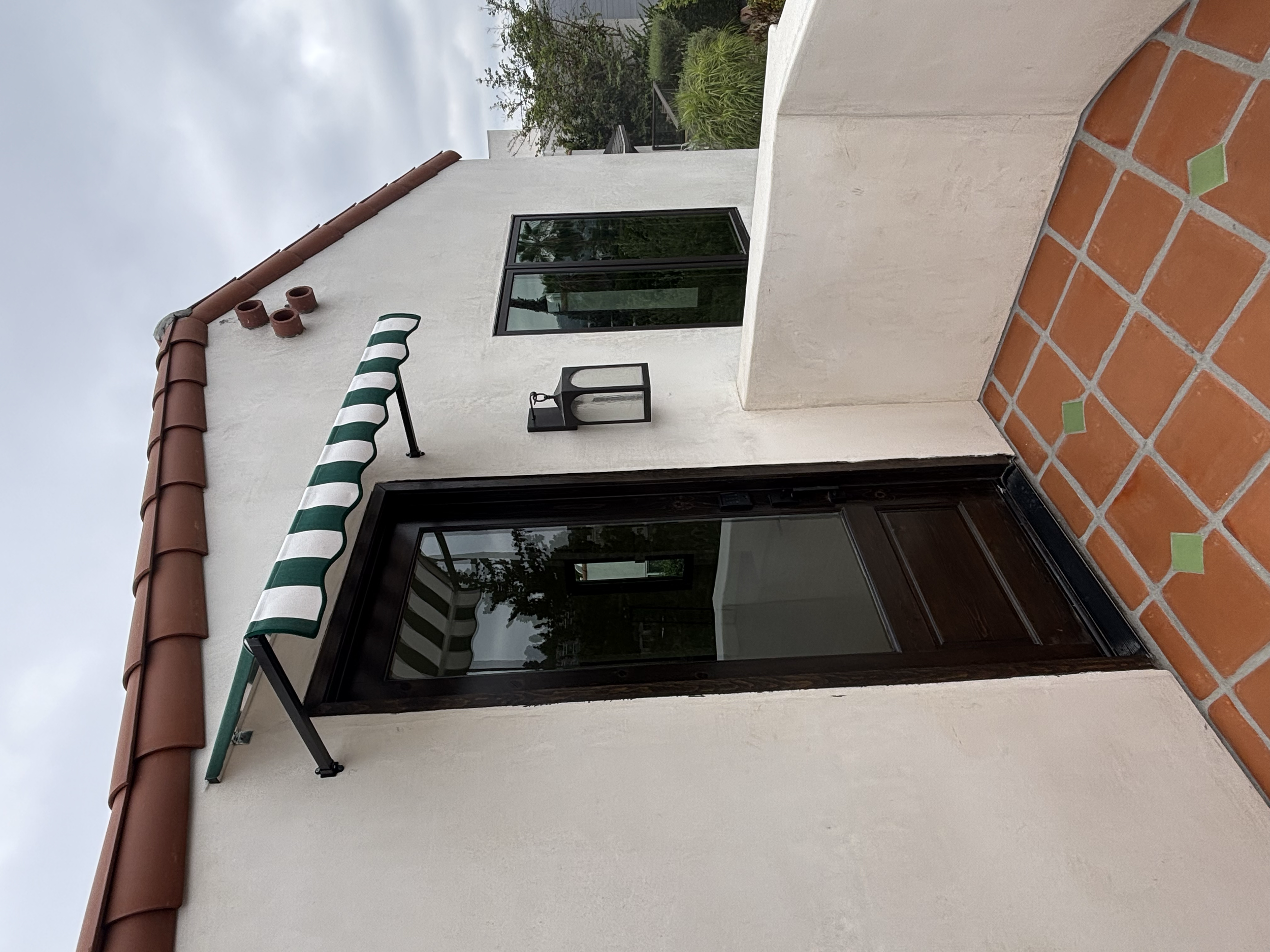 View of a balcony with a striped green and white awning, a black lantern-style light fixture, black-framed windows, beige stucco walls, and a terracotta tile floor. Greenery and trees are visible in the background under an overcast sky.