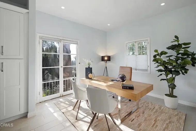 Bright home office with a wooden desk, beige office chair, white side chairs, large potted plant, window, glass door, and minimalist decor.