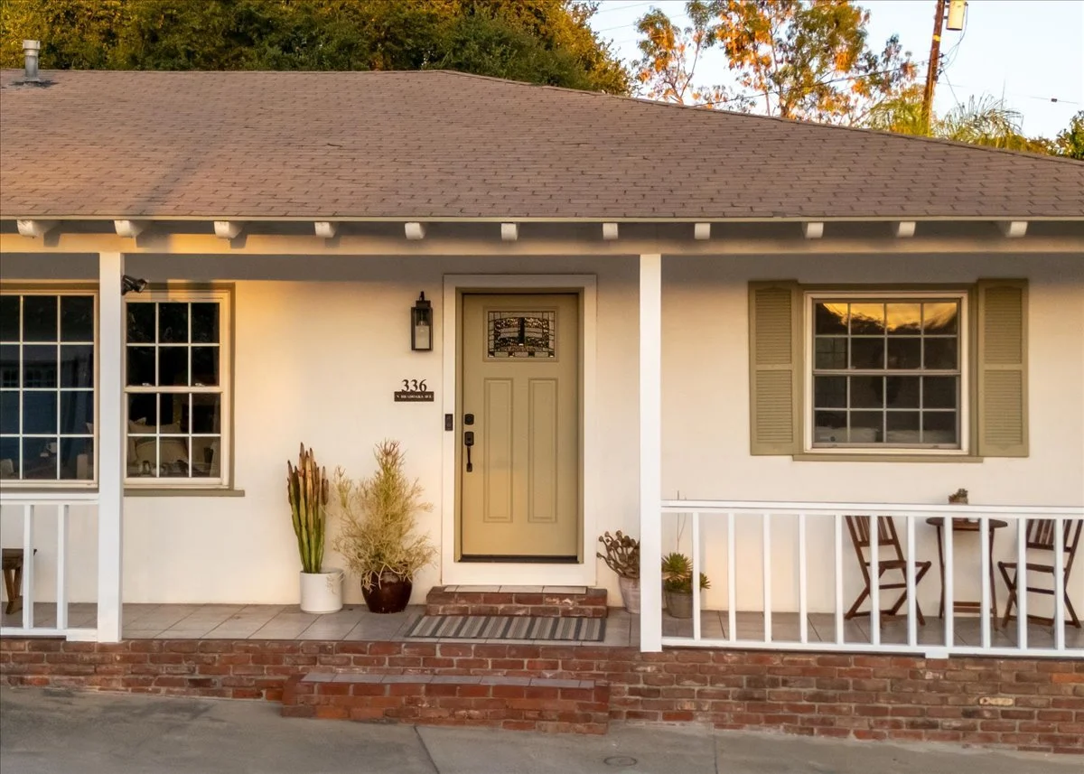 Front view of a house with a porch, a beige front door with a decorative glass window, surrounded by windows with beige shutters, potted plants near the entrance, and a brick step leading up to the door.