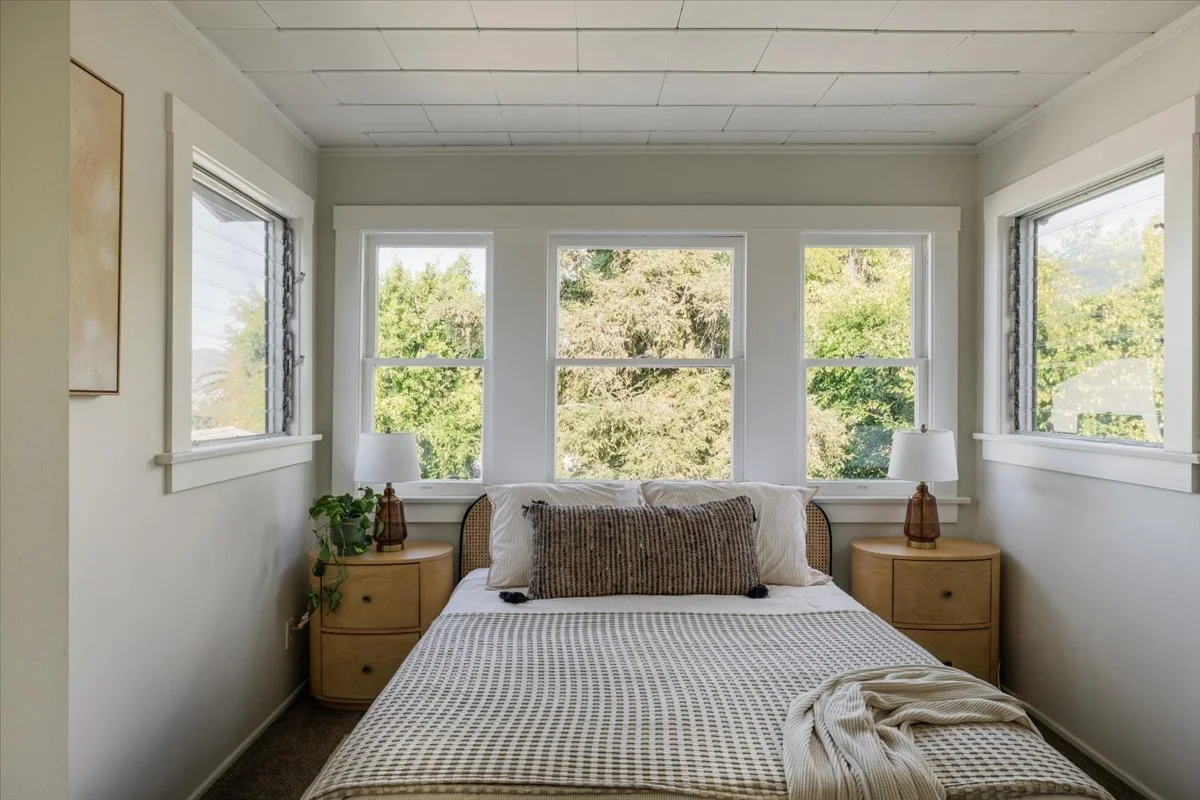 A cozy bedroom with a bed, pillows, nightstands, lamps, and three large windows showing greenery outside.