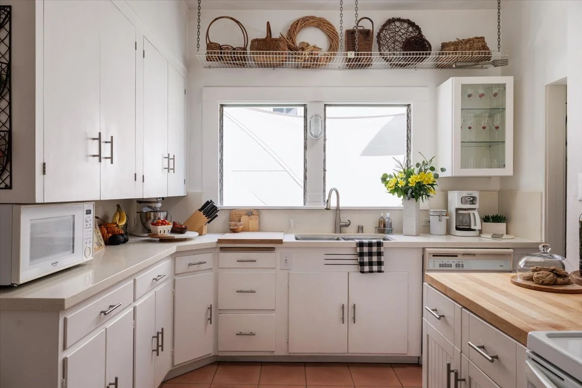 White kitchen with upper and lower cabinets, a countertop with a microwave, a bowl of fruit, and a knife block. A window above the sink shows outdoor light, and a vase with yellow flowers is on the counter. A breakfast bar with cookies under a glass 